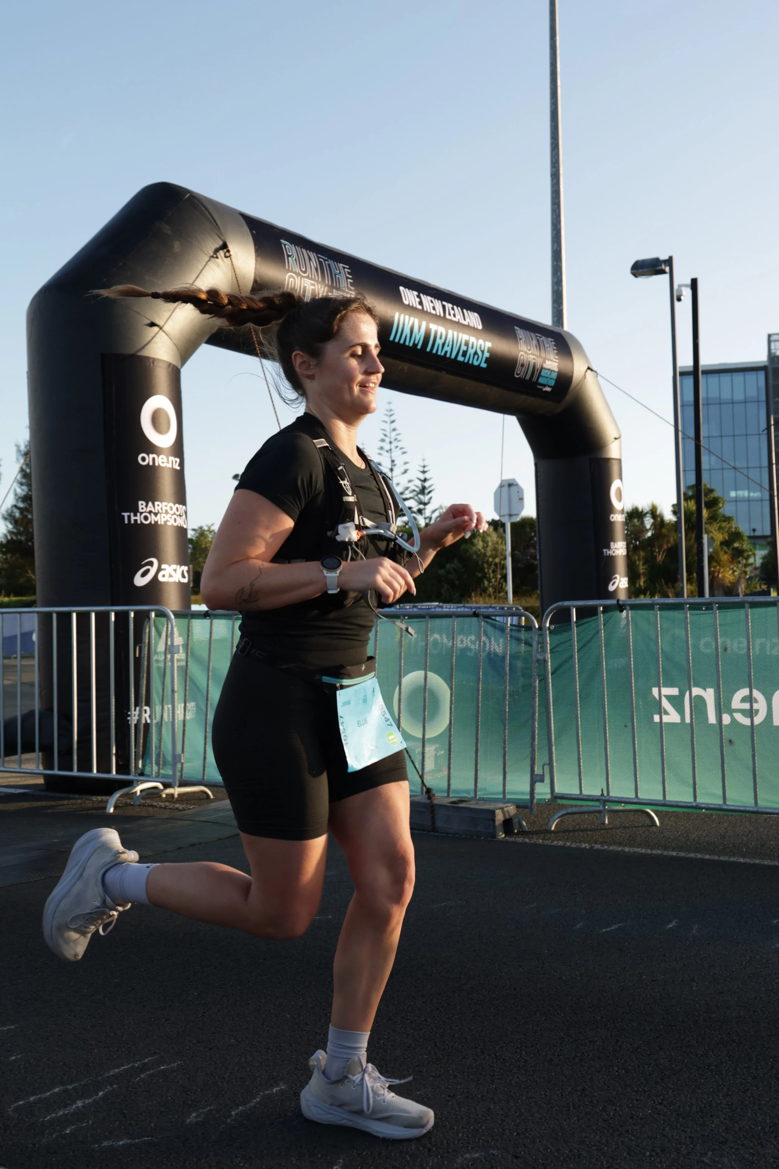 A woman running in a race, wearing athletic clothing and white sneakers, on a black track near race banners and barriers, with buildings and trees in the background.