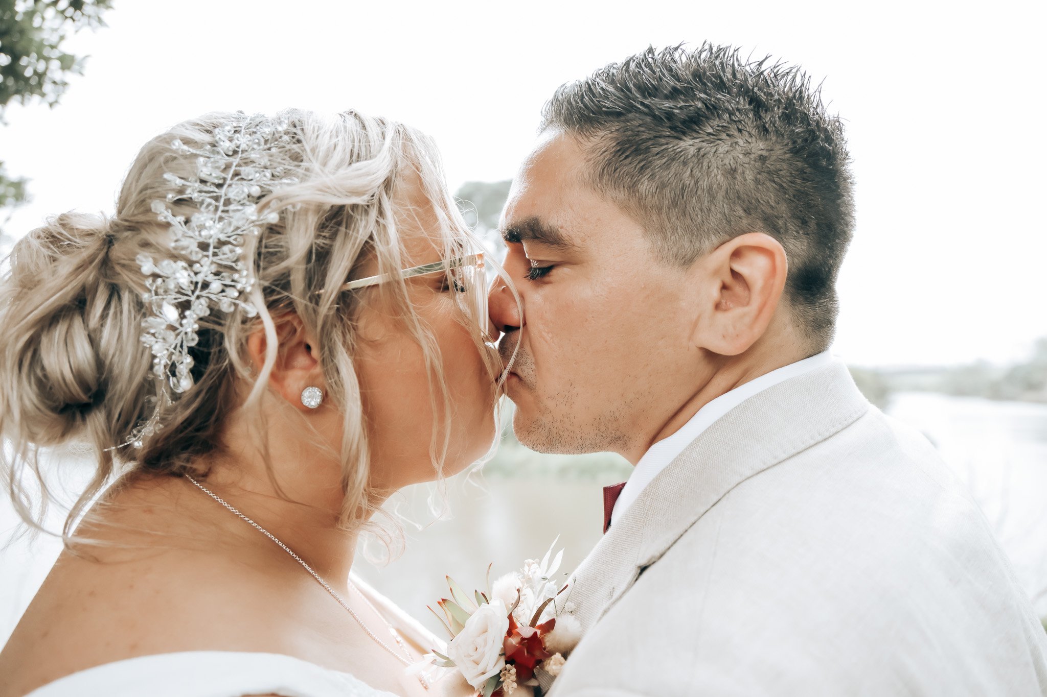 A bride with blonde hair styled in loose curls with decorative hairpins, wearing glasses, and a pearl necklace, and a groom with short dark hair and a white suit, sharing a close moment with eyes closed during a wedding.