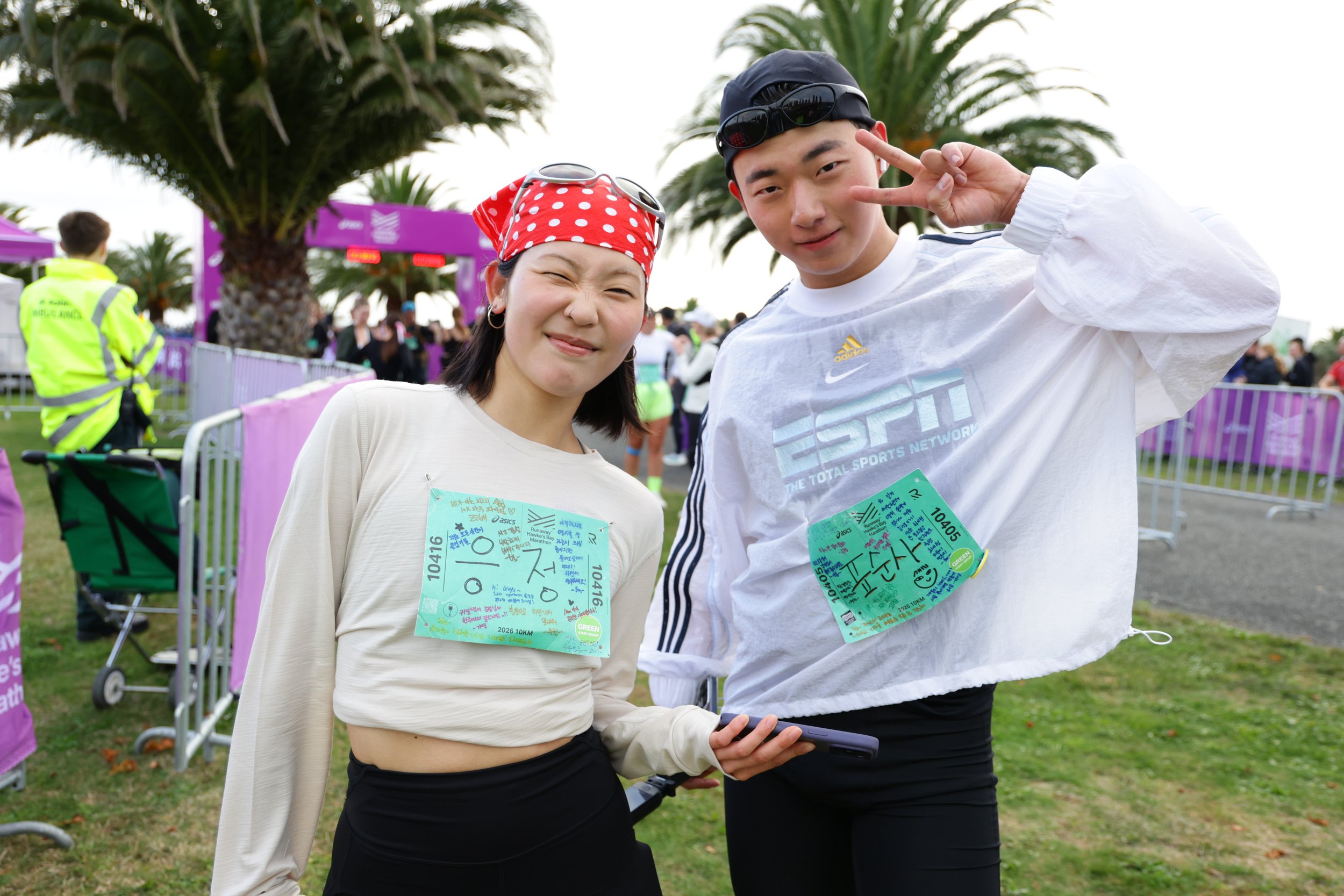 Two young runners, a woman with short dark hair in a white shirt with a red polka-dot headband, and a man with short black hair in a white Adidas shirt, posing for a photo at an outdoor event with palm trees and purple race banners in the background.