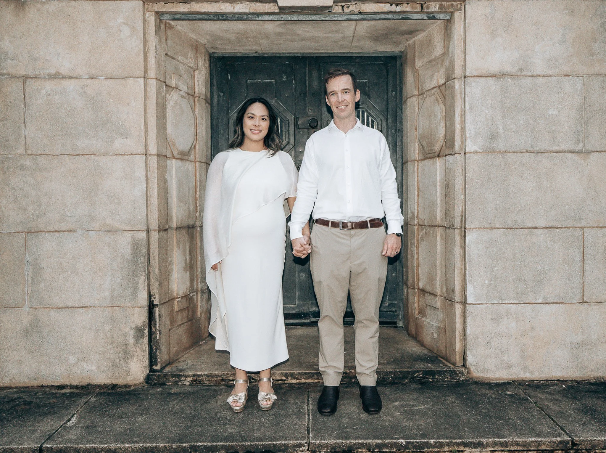 A couple standing in front of a stone entrance holding hands, dressed formally with the woman in a white dress and the man in a white shirt and beige pants.