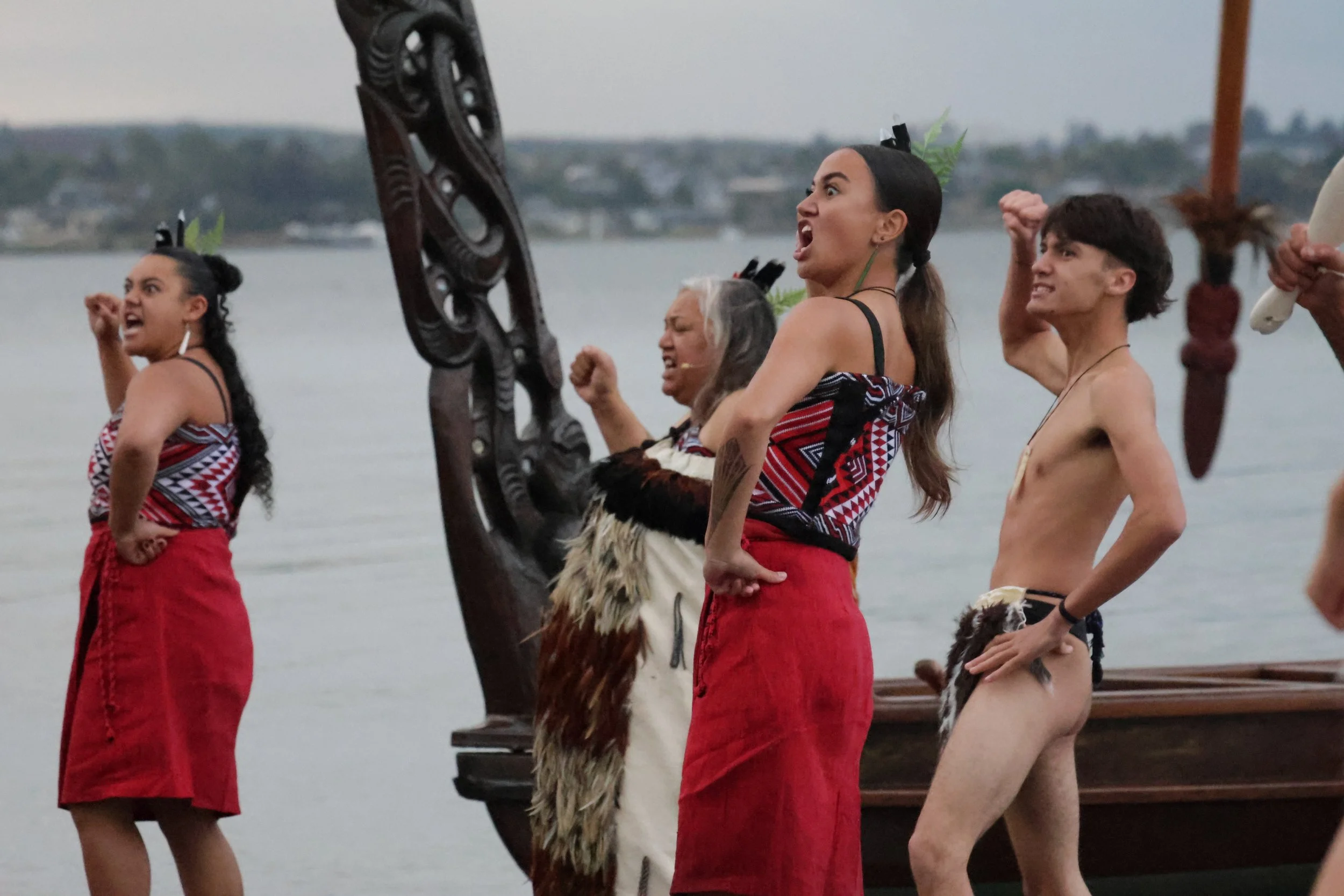 Group of indigenous people dressed in traditional clothing and body paint, standing in a row near a body of water, with some raising fists and others holding traditional items.