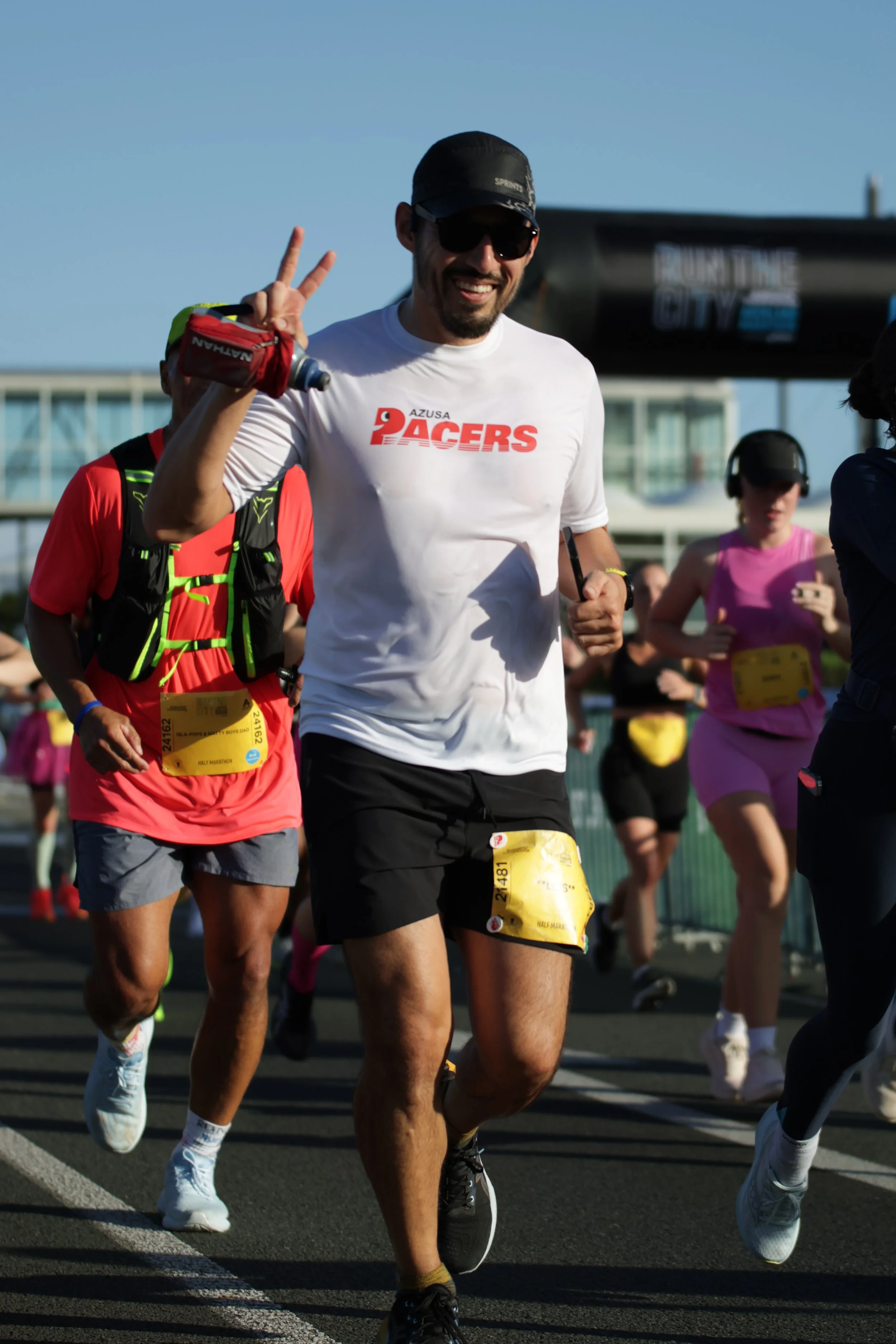 A group of marathon runners participating in a race, with a prominent man in a white "AZUSA PACERS" shirt making a peace sign and smiling, surrounded by others in colorful athletic wear, during daytime on a city street.