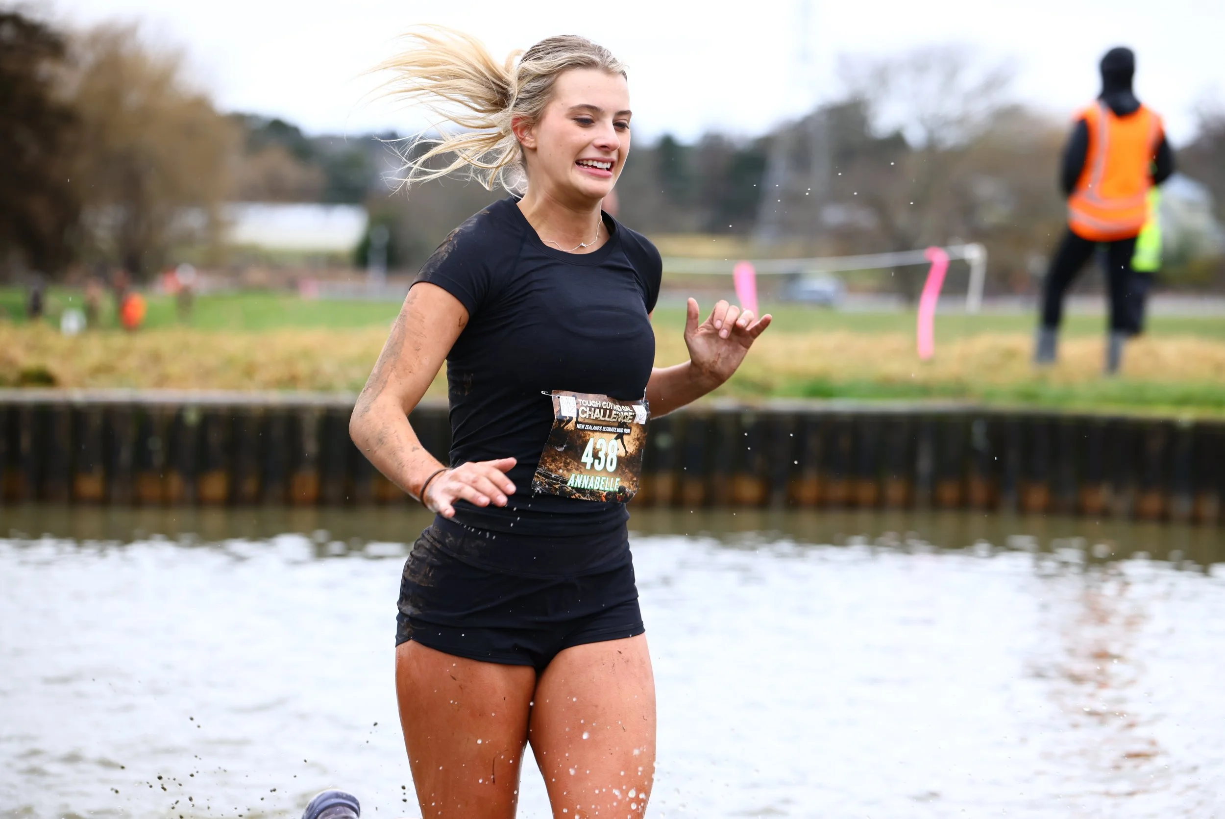 A woman running outdoors near water, with her hair in a ponytail, wearing a black athletic shirt and shorts, with a race bib number 438.