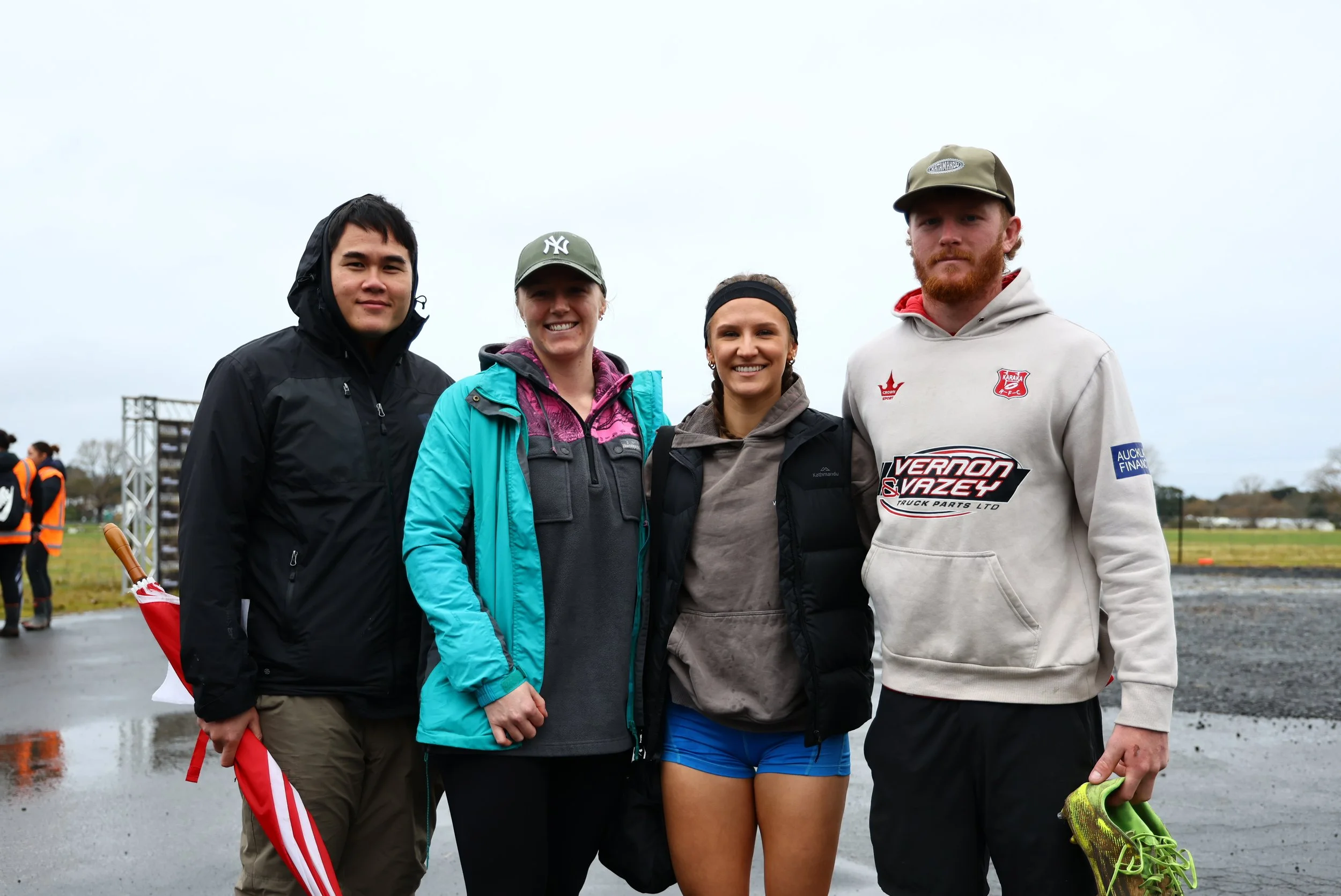 Four young adults, two men and two women, standing outdoors on a rainy day, smiling for the camera. They are dressed in casual and athletic clothing, with some holding sports gear and shoes.