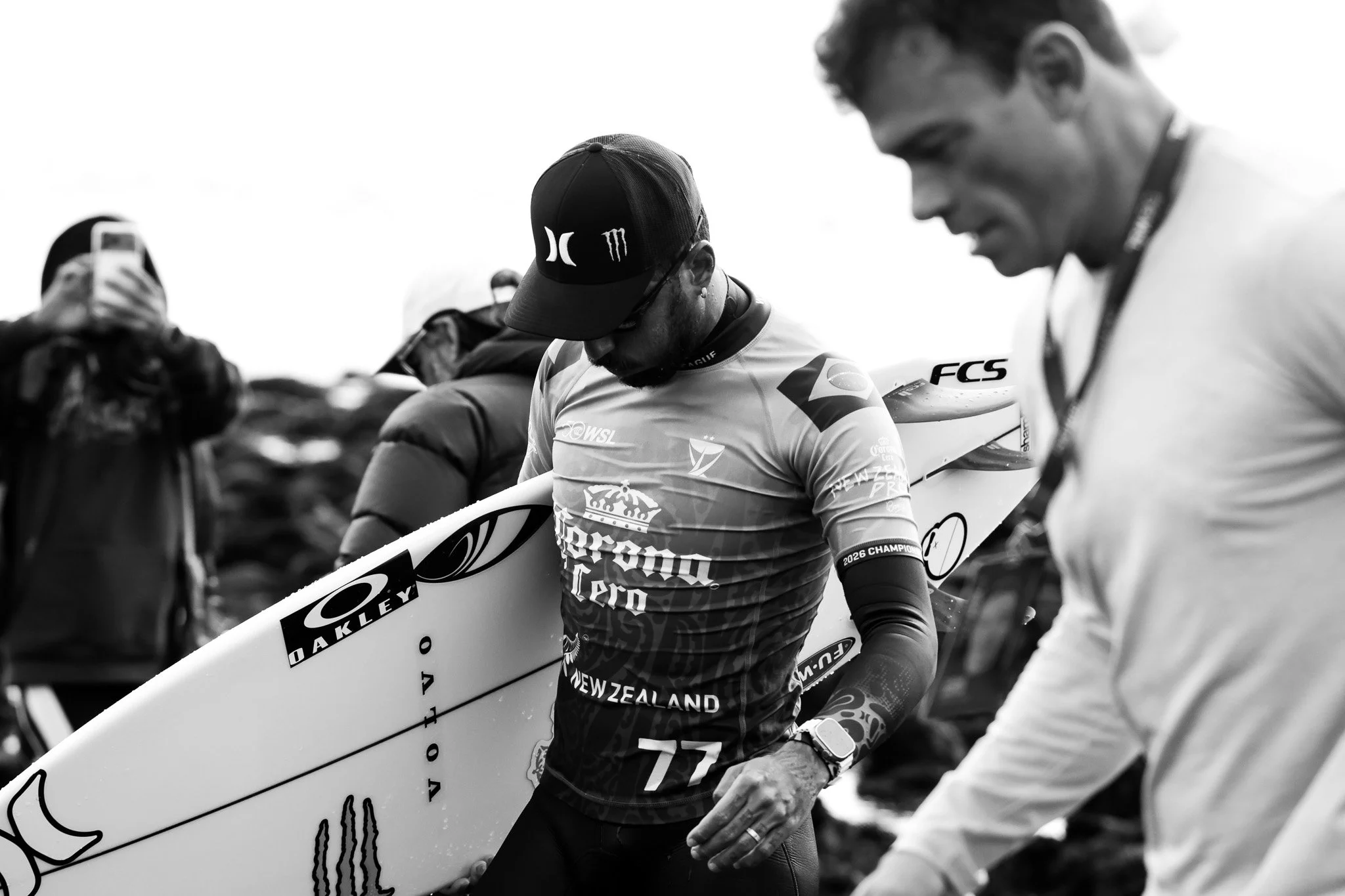 Black and white photo of athletes preparing for a race, with one holding a surfboard-like racing board, wearing racing gear with sponsors' logos, and surrounded by other team members.