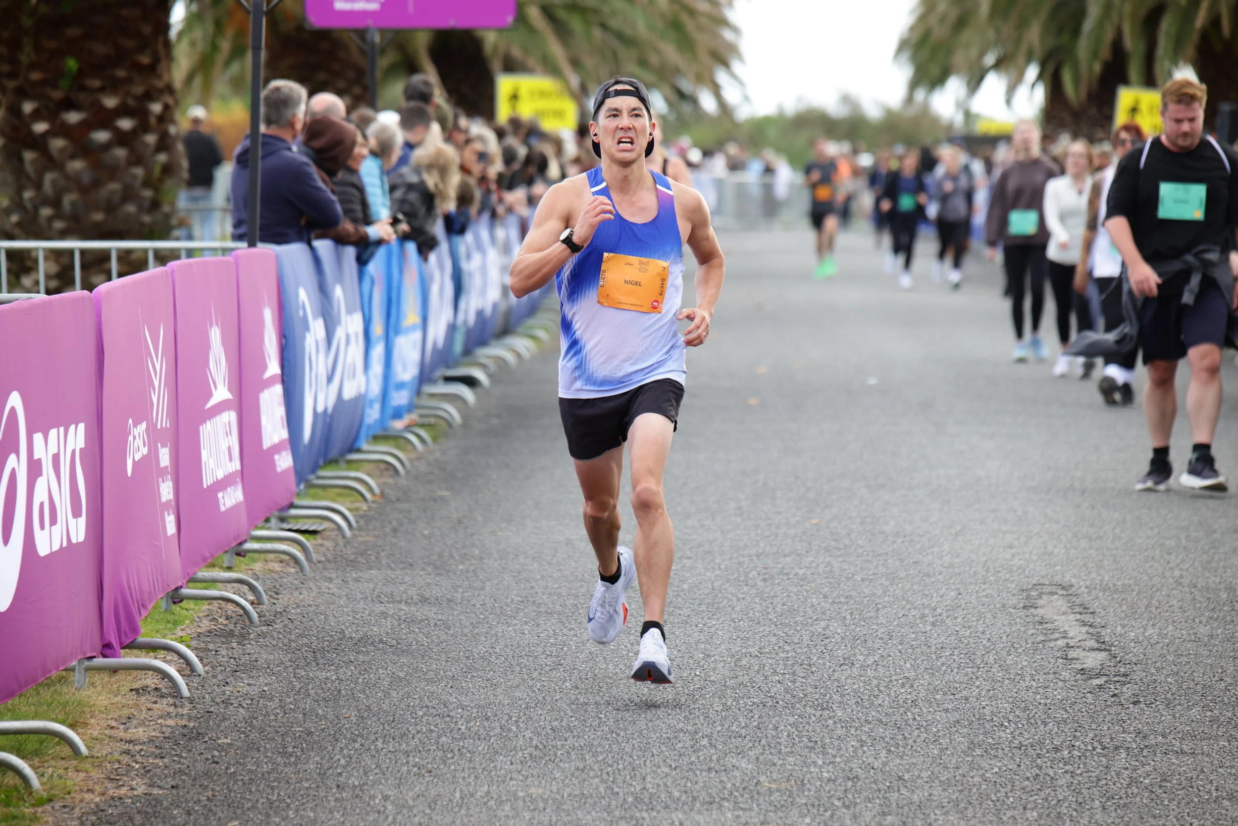 A man running in a race, wearing a blue tank top, black shorts, a black cap backwards, and a watch, with a crowd of spectators behind purple barricades and trees in the background.