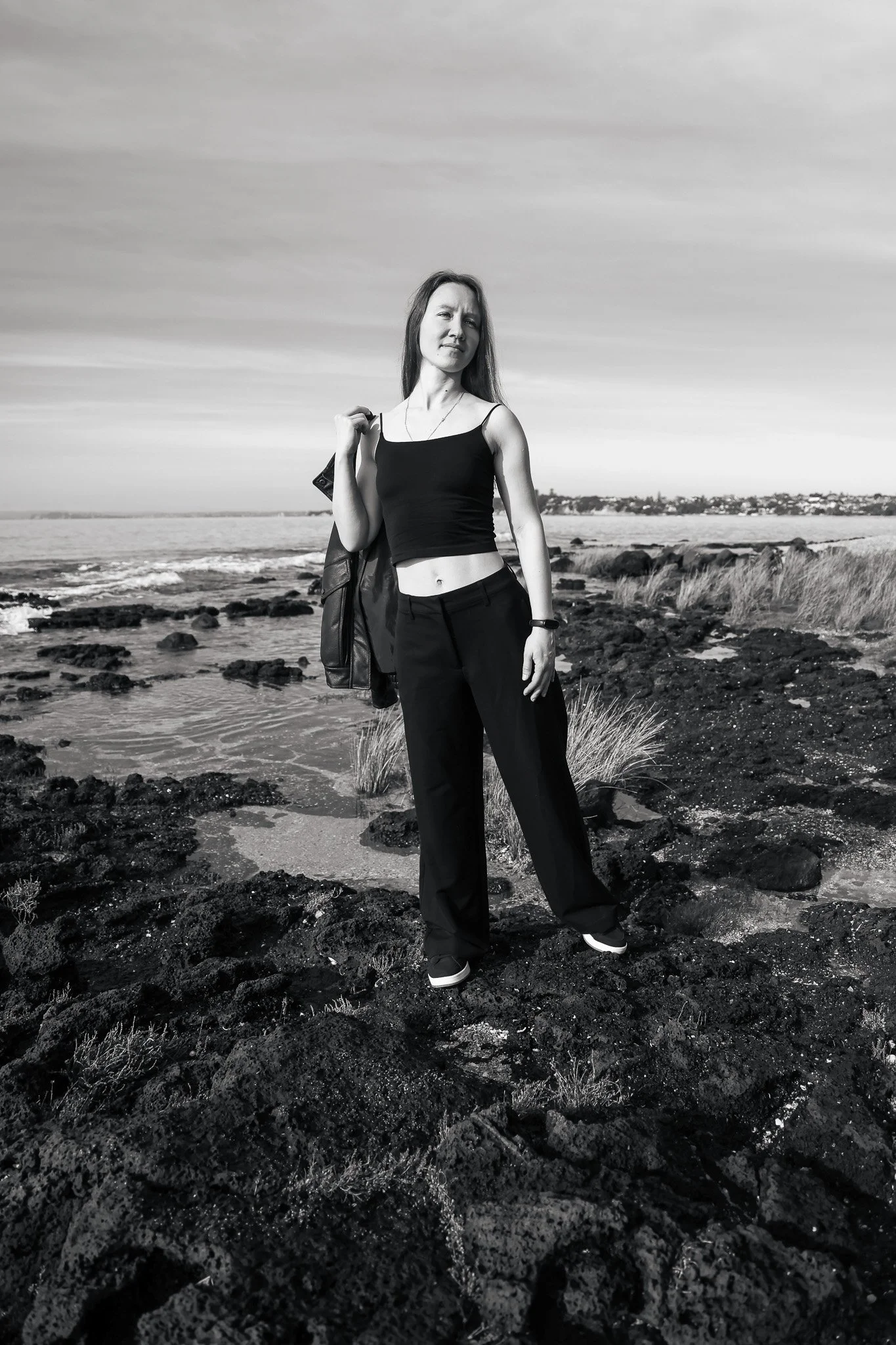 A woman standing on a rocky shoreline near the ocean, wearing black pants and a black crop top, holding a jacket over her shoulder, with a distant view of houses along the coast and a cloudy sky.