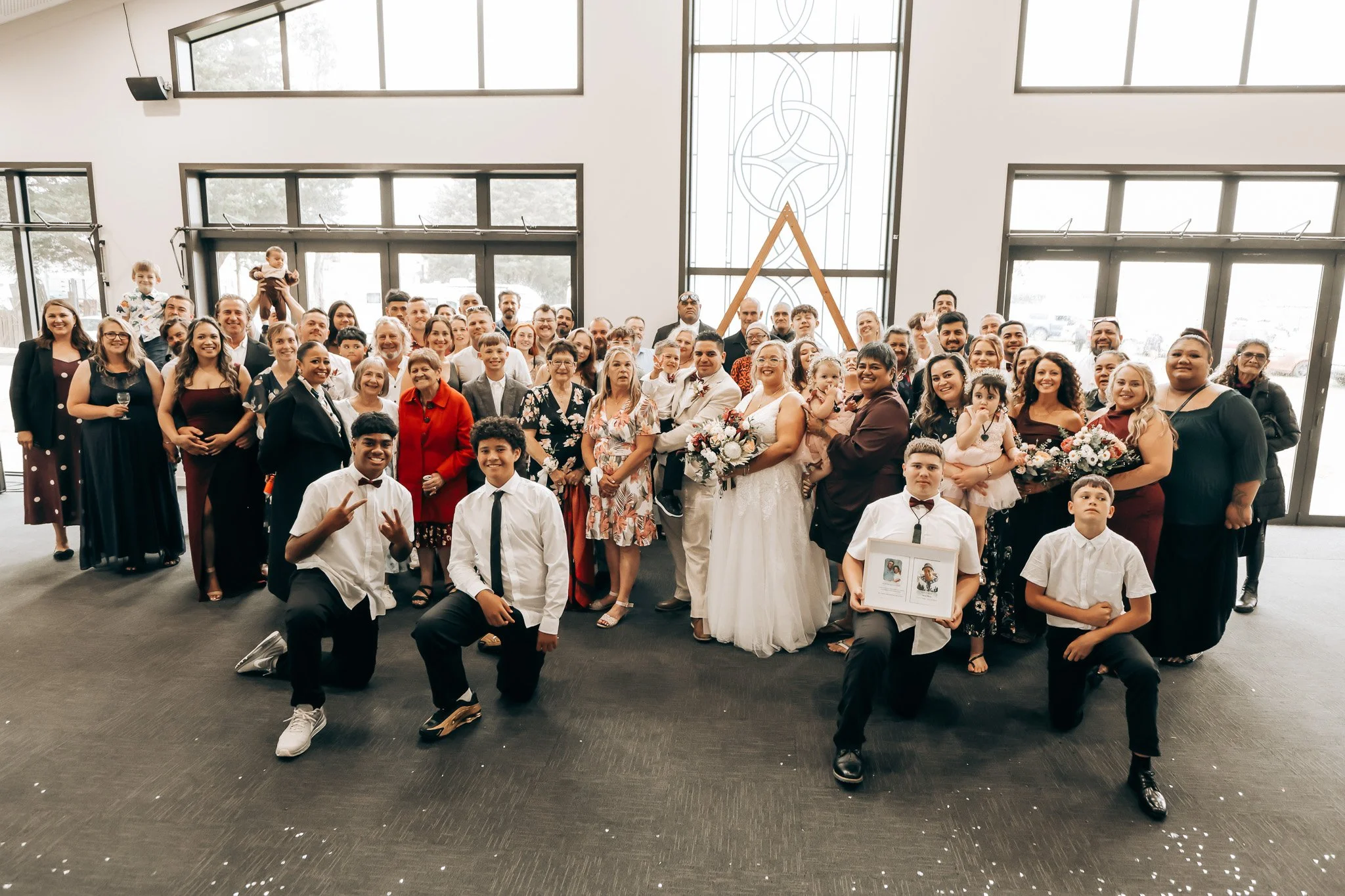 A large group of diverse people gathered inside a modern church, celebrating a wedding with several children present. The bride and groom are in the center, smiling, with the bride holding a bouquet. The group includes men, women, and children of var