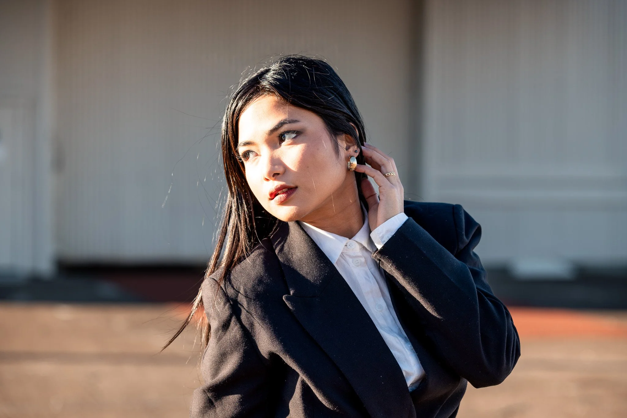 A young woman with black hair styled straight, wearing a black blazer and white blouse, standing outdoors with sunlight on her face, touching her ear with her left hand, against a blurred background of a building and pavement.