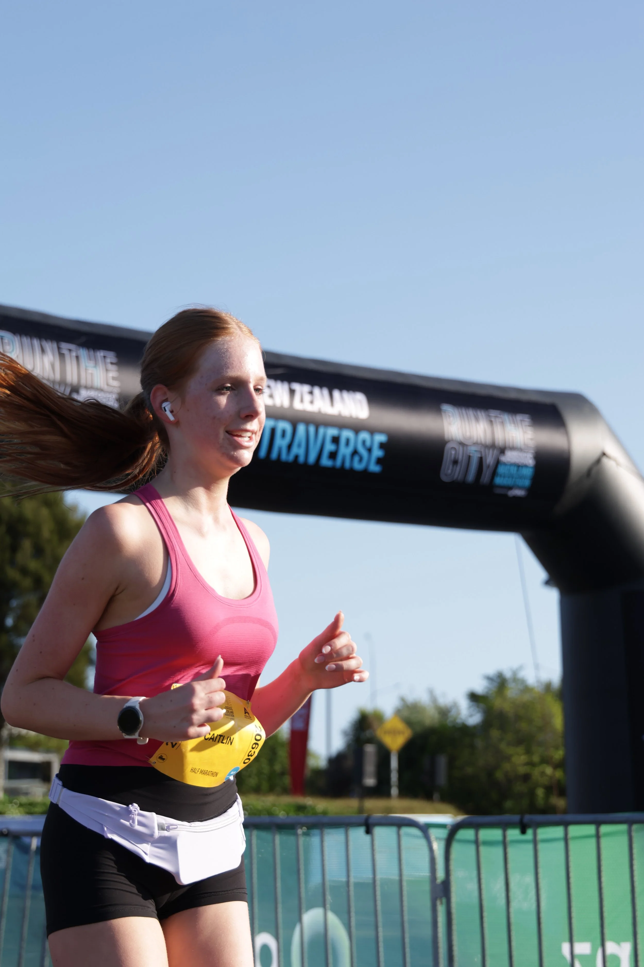 A young woman is running in a race, wearing a pink tank top, white and black shorts, a bib number, and wireless earbuds, with a race arch and trees in the background on a sunny day.