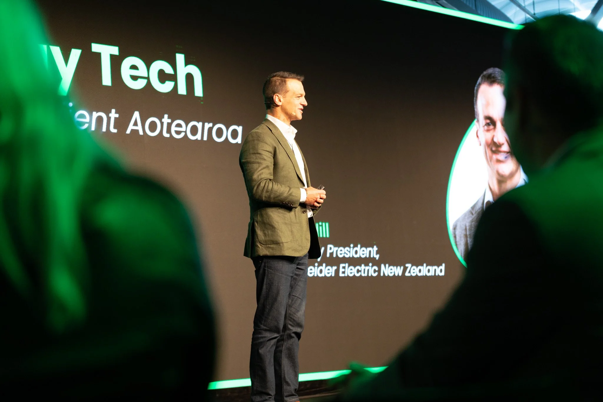 A man in a suit on stage at a tech conference, speaking with a large screen behind him displaying his name and title. The screen reads "Y Tech, President Aotearoa" and mentions "Seider Electric New Zealand." Green lighting and blurred audience member