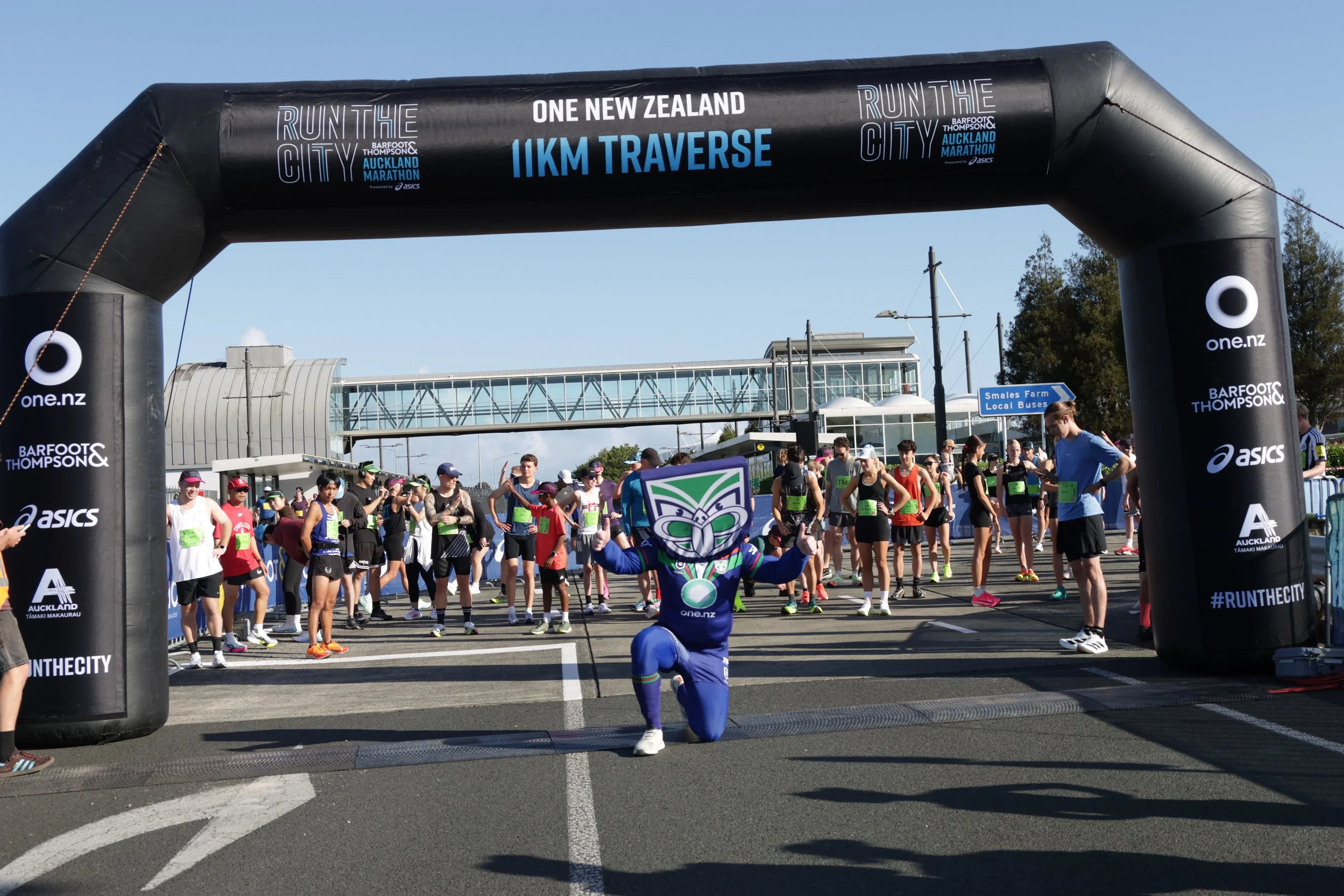 Participants at the starting line of a running race, with some smiling and dressed in athletic gear, under a large black inflatable arch that reads 'Run the City Auckland Marathon' and '1KM Traverse'. The event is sponsored by Asics and Barfoot & Tho