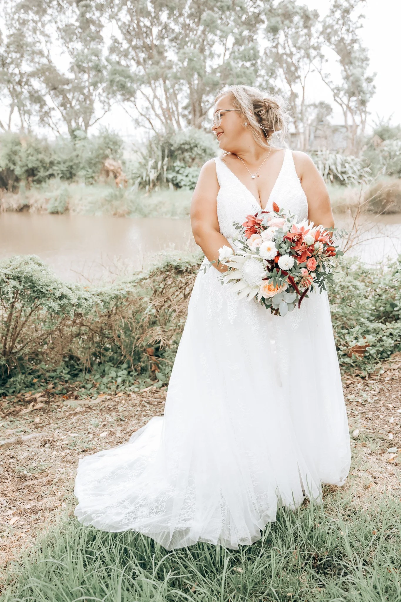 A woman wearing a white wedding dress and glasses holding a bouquet of flowers outdoors near a body of water, with trees and greenery in the background.