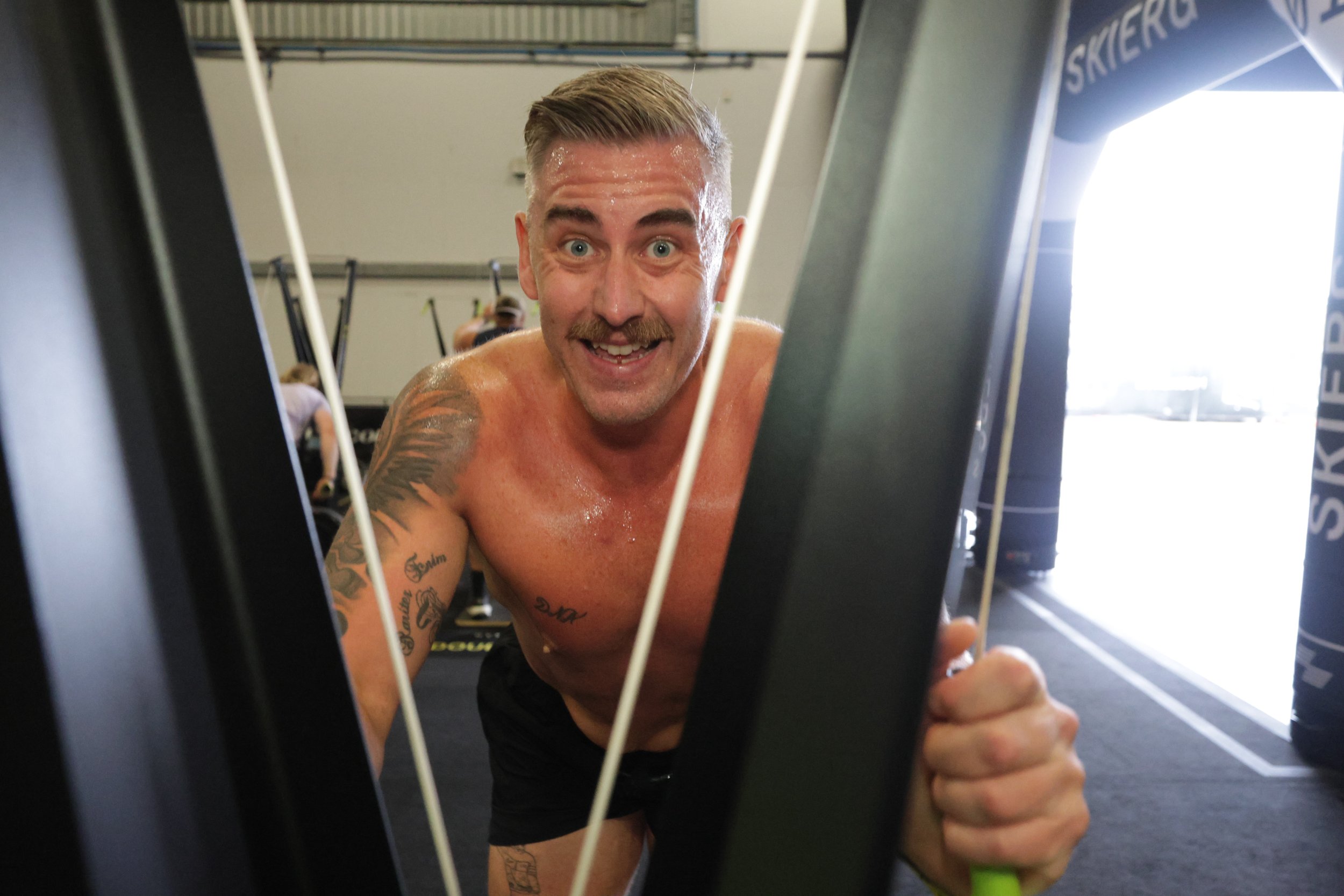 A shirtless man with tattoos on his arm and chest, working out on a treadmill in a gym, smiling and sweaty, looking through the treadmill's frame.