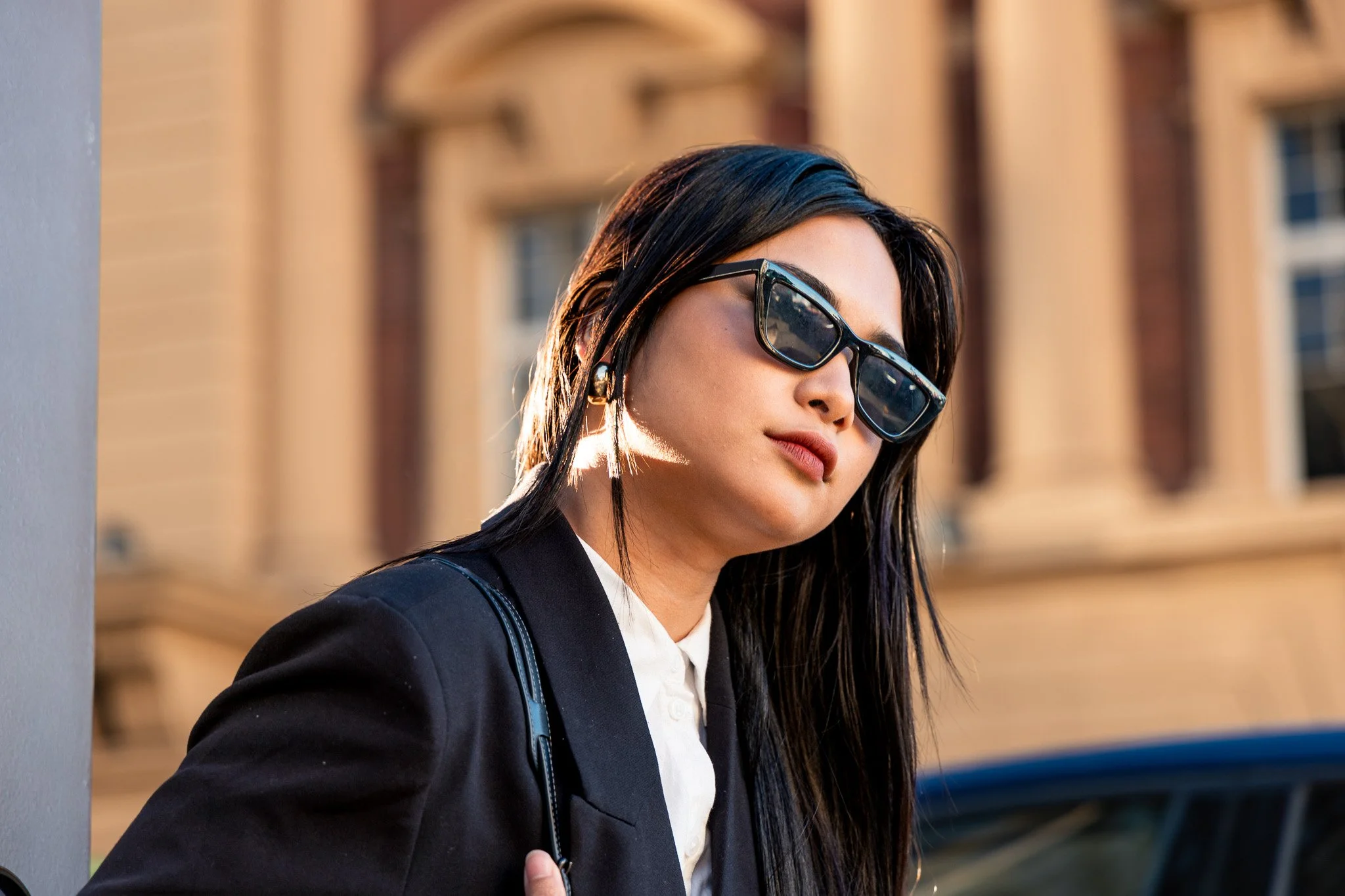 Woman with long dark hair wearing black sunglasses, black blazer, and white shirt, standing outdoors in front of a brick building.