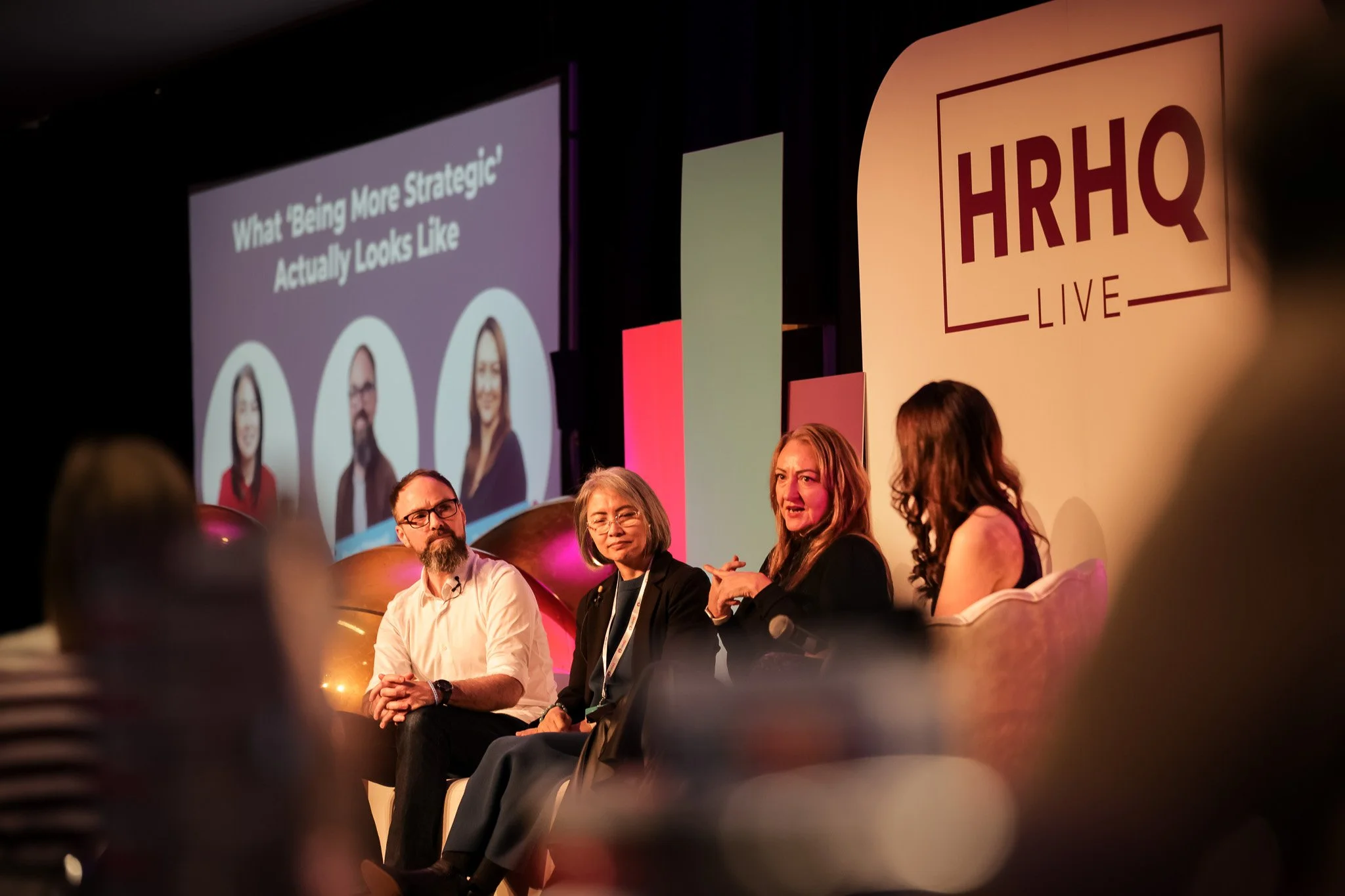 A panel discussion at HR & Q Live conference with four people sitting on stage, a large screen displaying a presentation slide with portraits of three women and the title 'What Being More Strategic Actually Looks Like', and a large HR & Q Live sign o