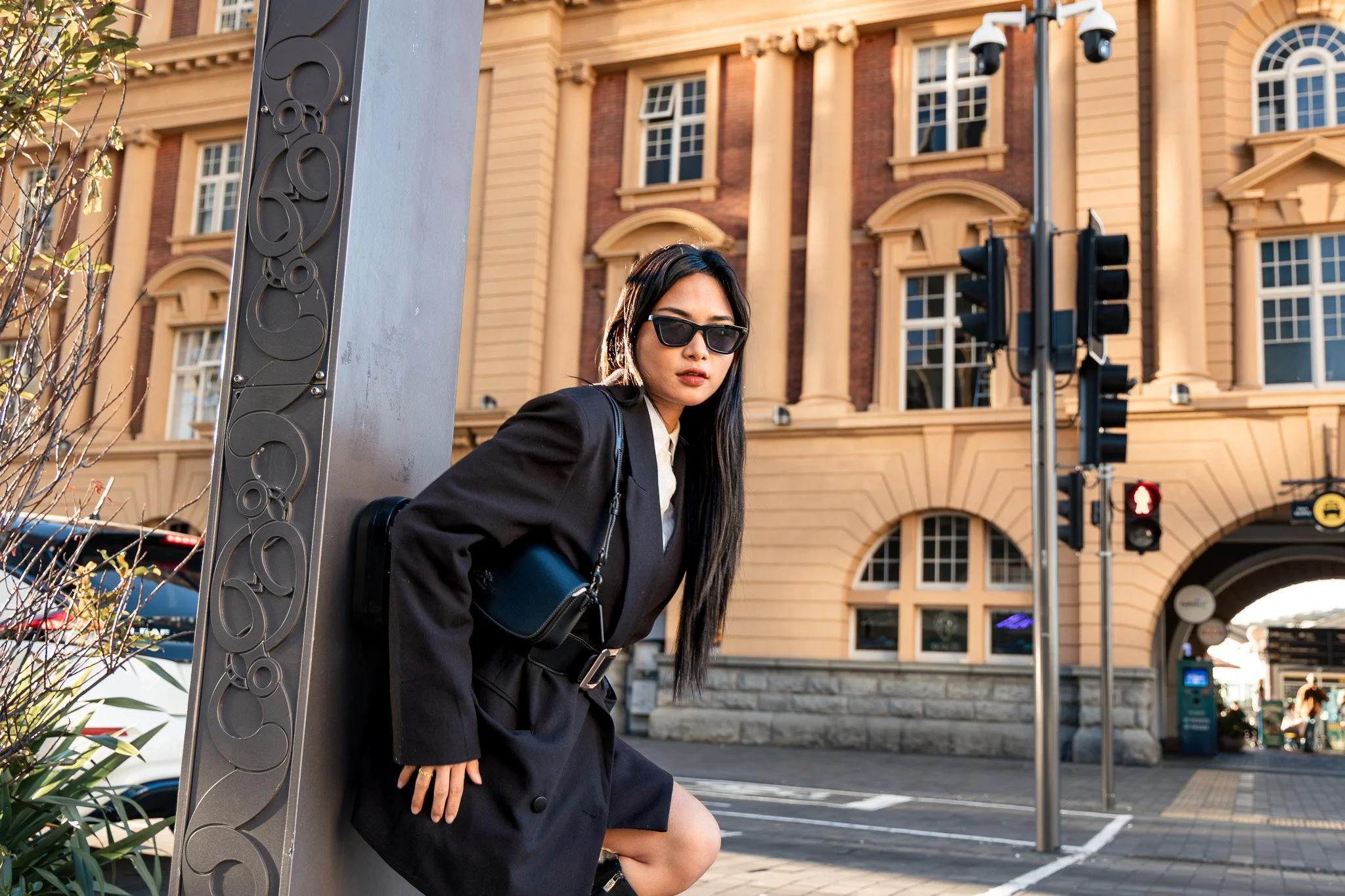 A young woman with long dark hair and sunglasses is sitting on a city street corner, leaning against a decorative pole with traffic lights behind her. She is wearing a black blazer and a white shirt, with a black handbag over her shoulder. The backgr