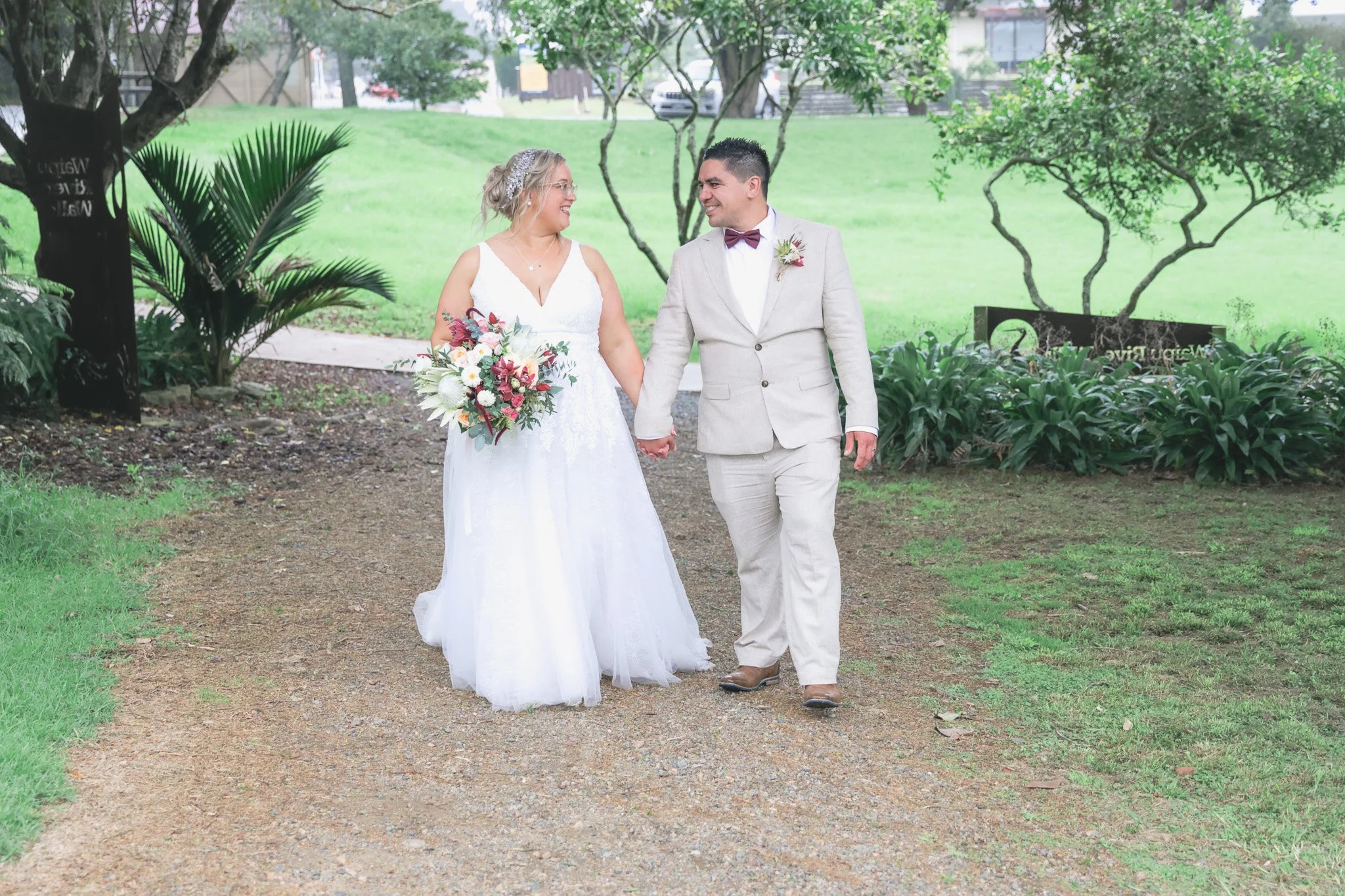 A bride and groom walking hand in hand outdoors in a park, smiling at each other. The bride wears a white wedding dress and holds a bouquet, while the groom wears a light-colored suit with a bow tie. The background features greenery and trees.