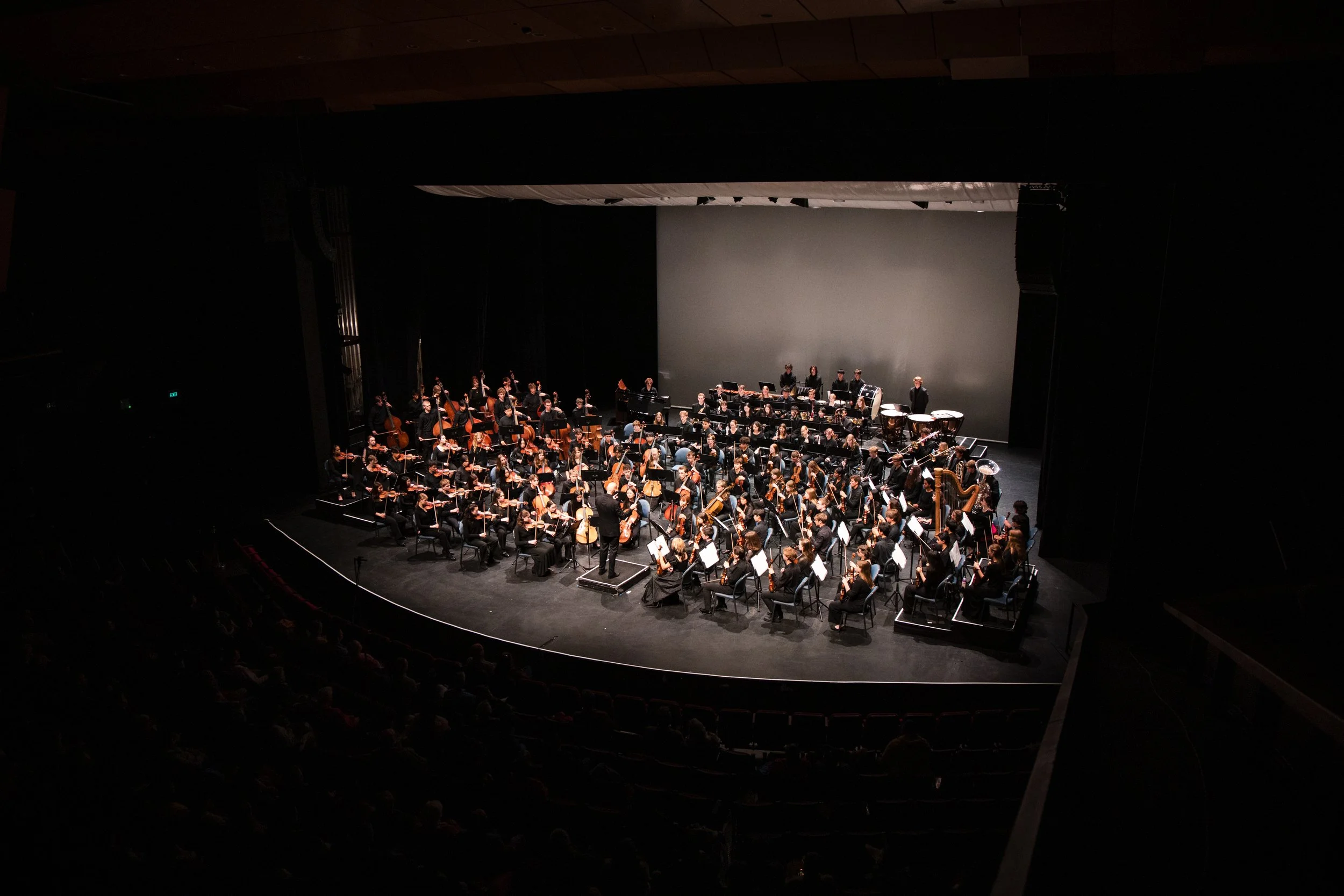 An orchestra performing on stage in a concert hall with a large gray backdrop. The musicians are seated with their instruments, and a conductor is standing in front of them. The stage is well-lit, and the audience seating is visible in the darkness.