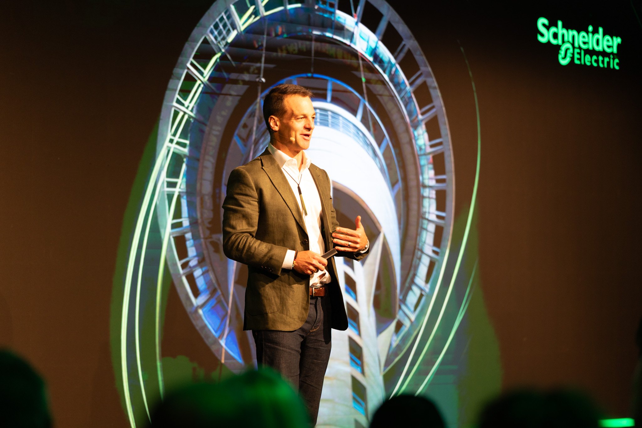A man in a suit giving a presentation on a stage with a large digital display behind him. The display shows a green logo for Schneider Electric and an abstract circular graphic in vibrant colors.