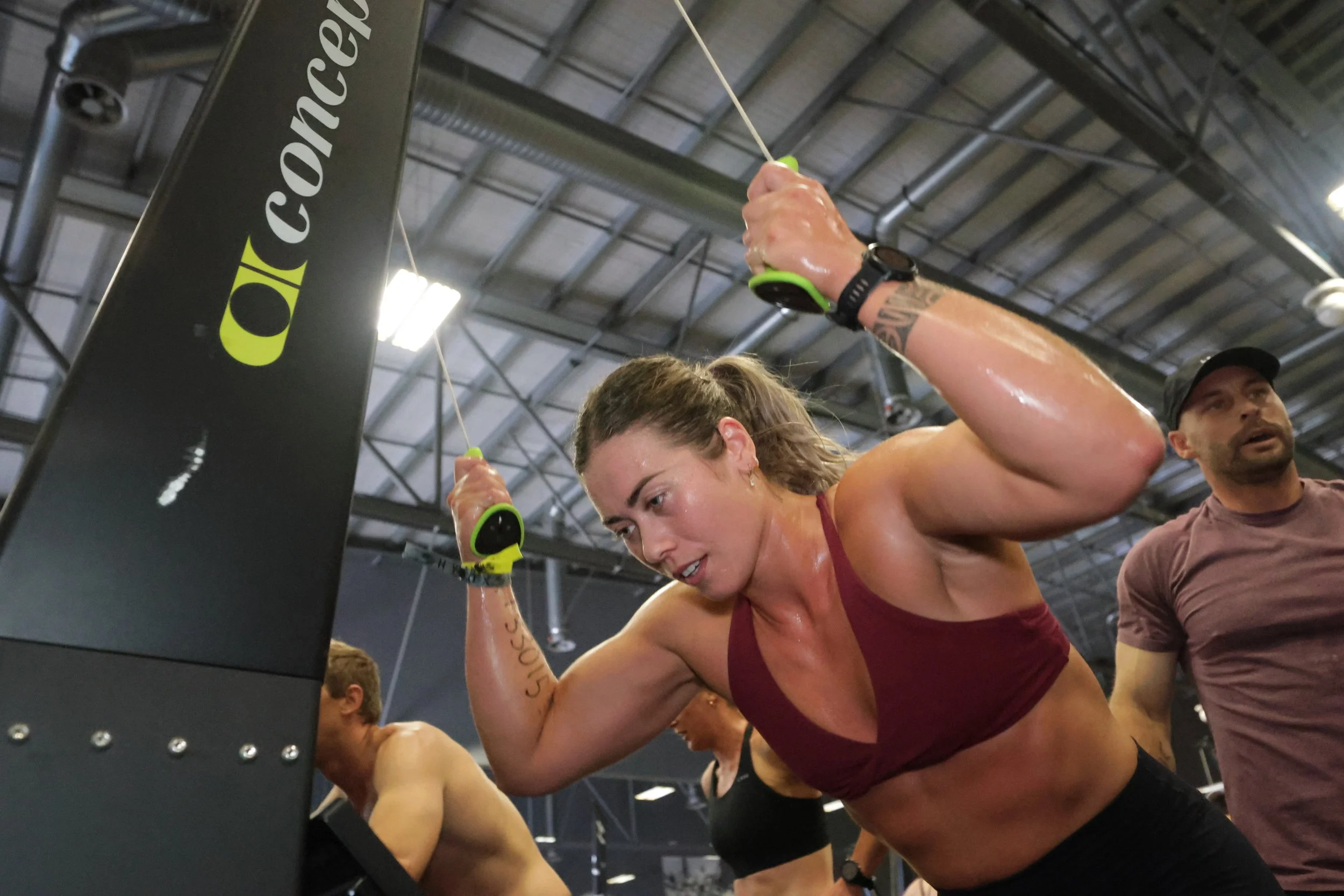 A woman with a maroon sports bra and black workout pants is doing push-ups on a gym floor, holding onto ropes attached to a fitness machine.