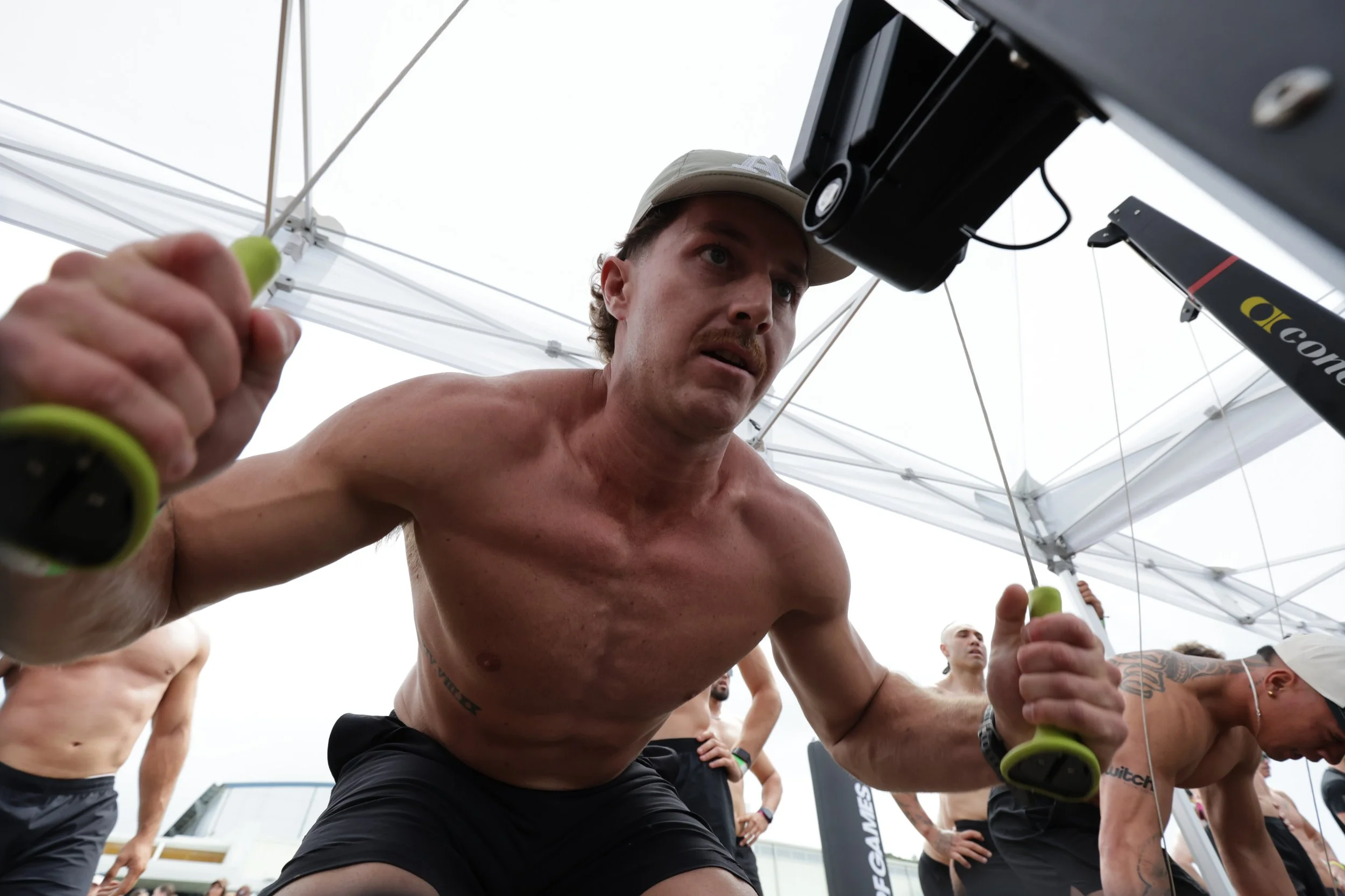 A shirtless man with a hat is participating in a fitness activity under a white canopy tent, surrounded by other shirtless men, while holding green handles, possibly during a workout session.