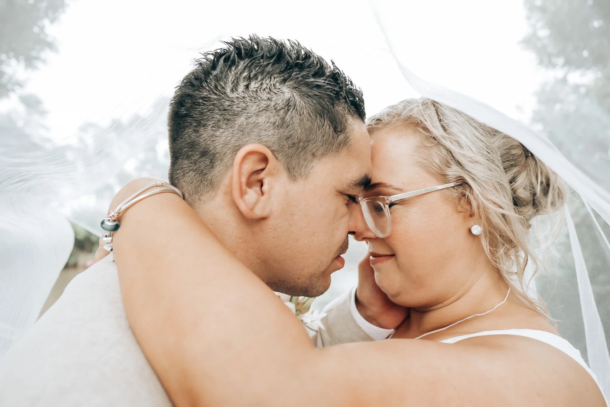 A close-up of a couple sharing an intimate moment with their foreheads touching and eyes closed, under a sheer white veil, outdoors with trees in the background.