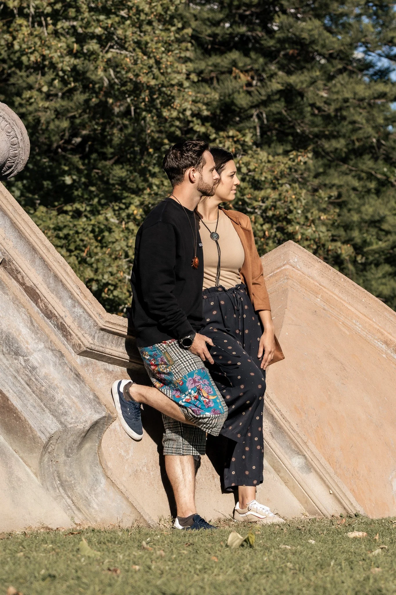 A young man kneeling in front of a young woman on a stone staircase outdoors with green trees in the background.