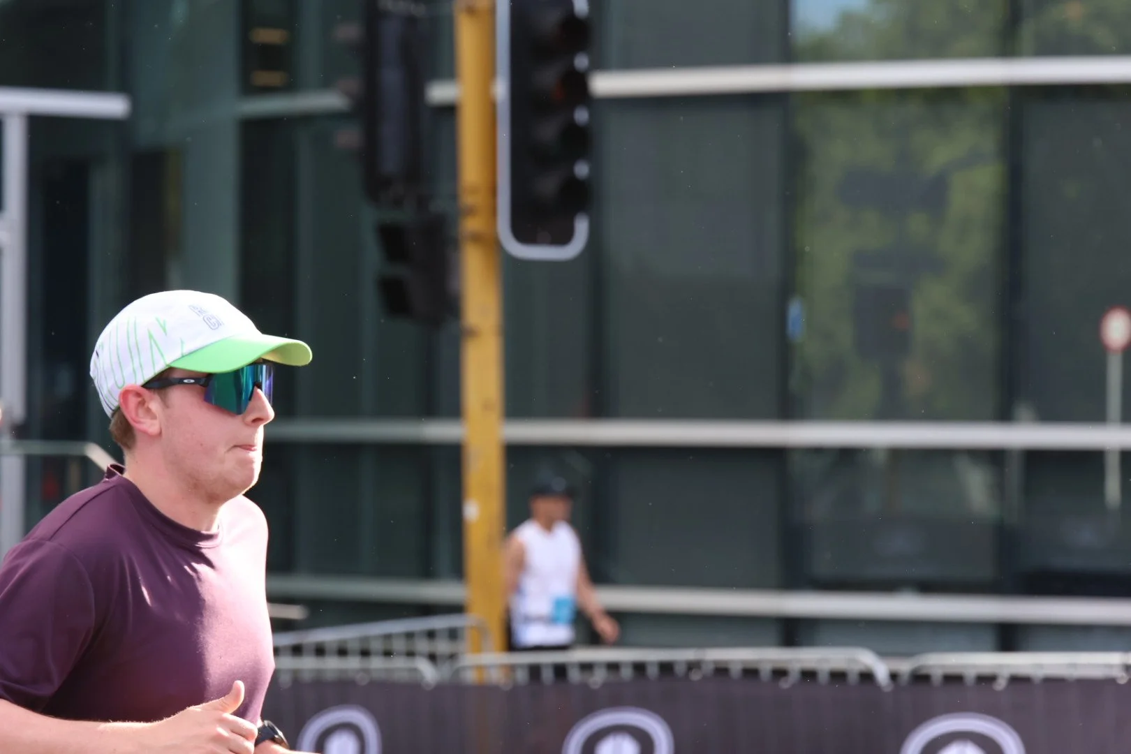 A person running outdoors, wearing a white cap with green accents, sunglasses, and a maroon shirt, with a street and traffic light in the background.