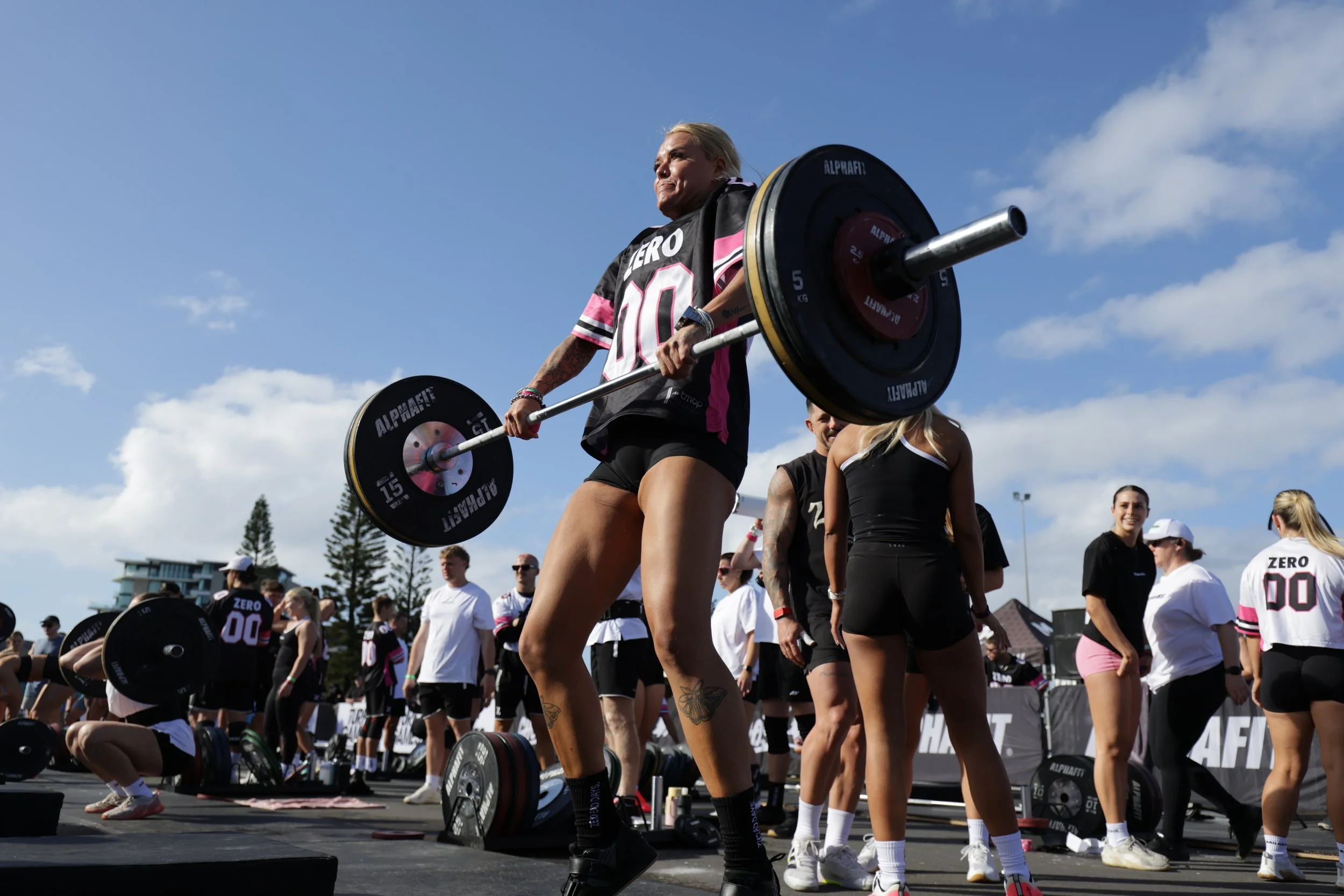 A woman lifting a barbell during a CrossFit competition outdoors under a blue sky, surrounded by other athletes and spectators.