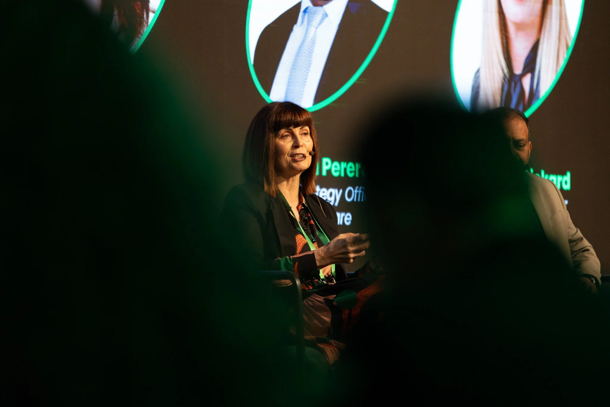 A woman with brown hair and a black blazer speaking at a conference or event with a microphone headset. She is sitting beside another person on a stage, with a large screen behind her displaying images of two people and text. The audience is partiall