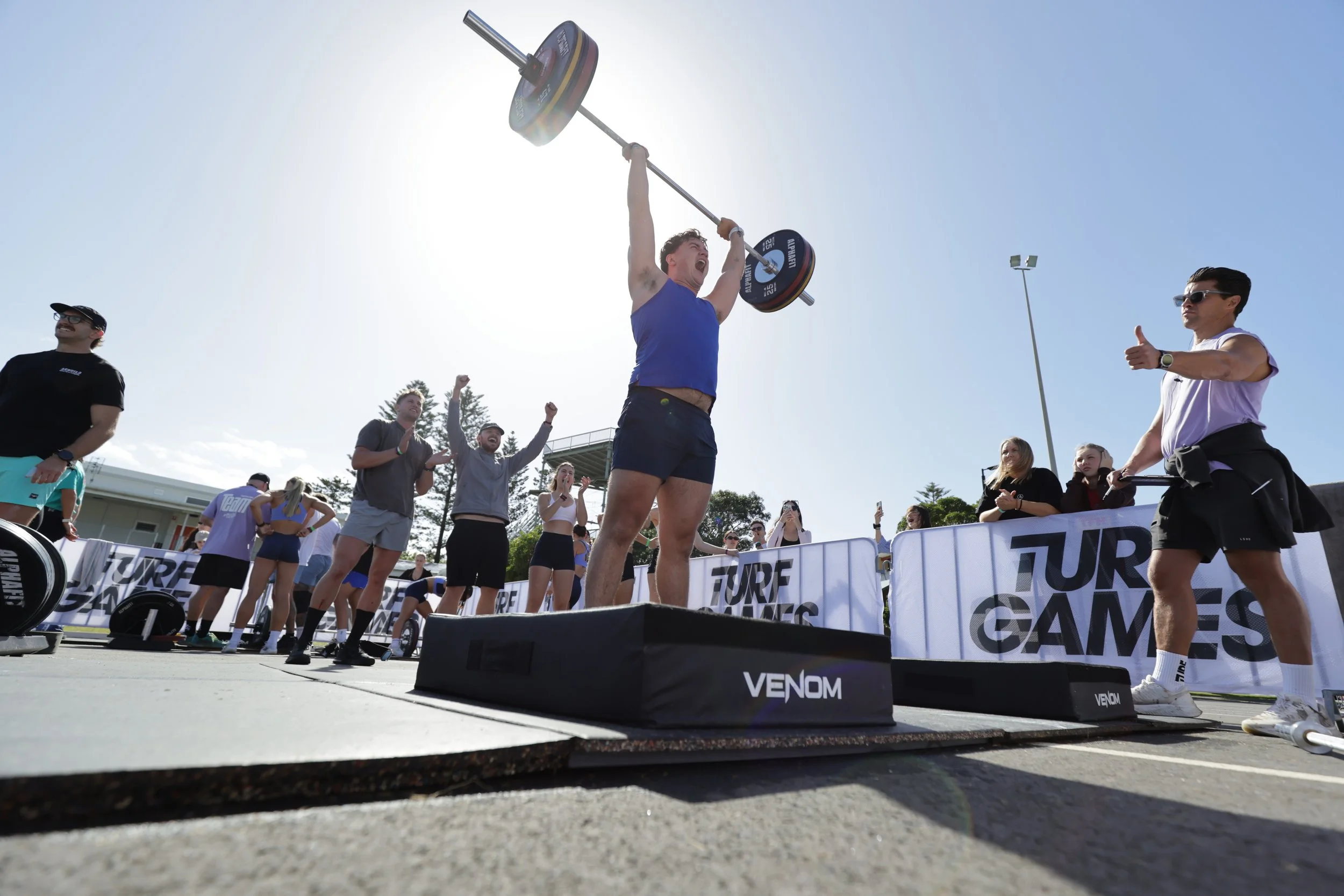 A man lifting a barbell overhead during a fitness competition with cheering spectators around him.