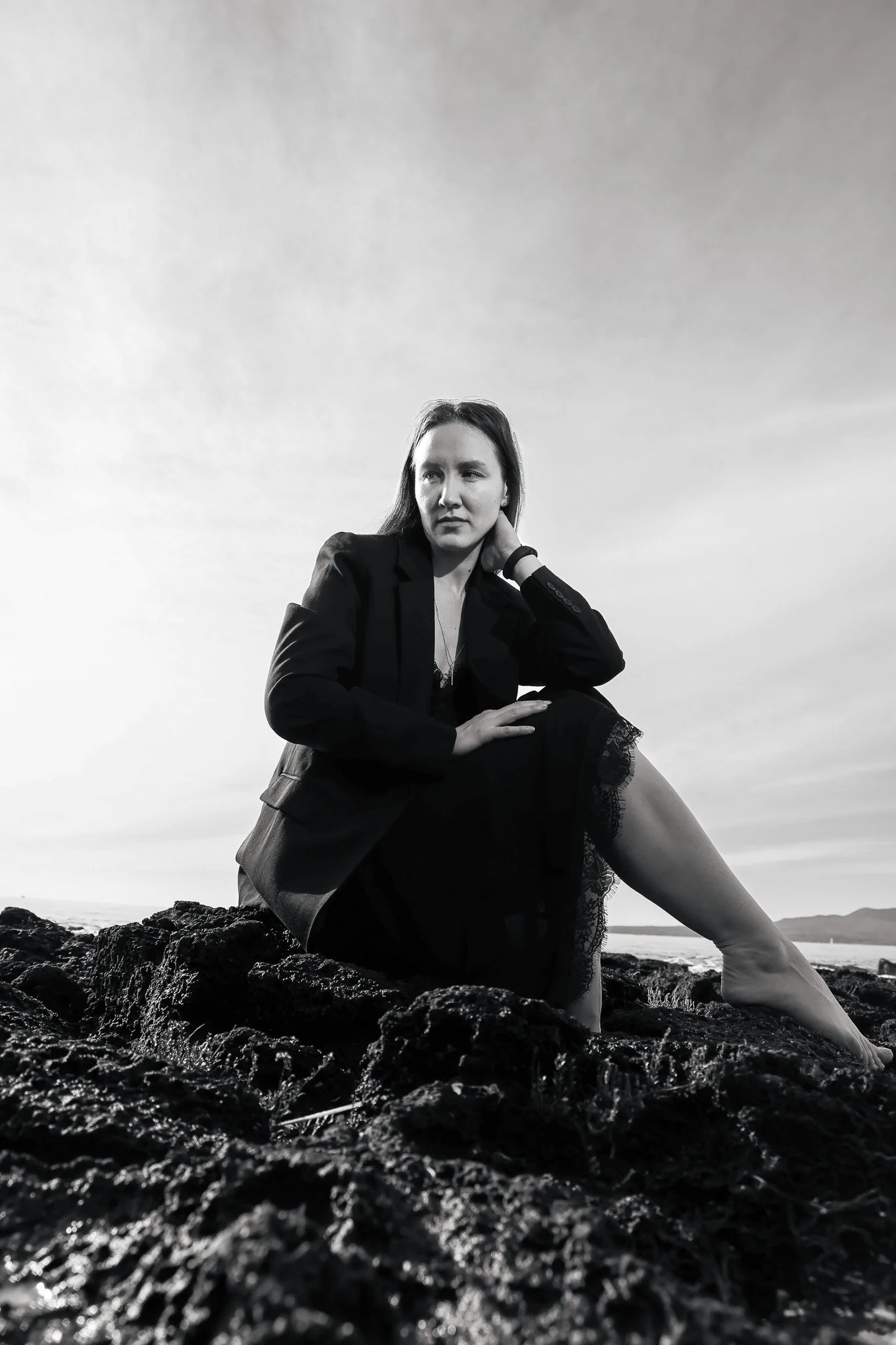 Black and white photo of a woman sitting on rocks near the sea, with a cloudy sky in the background