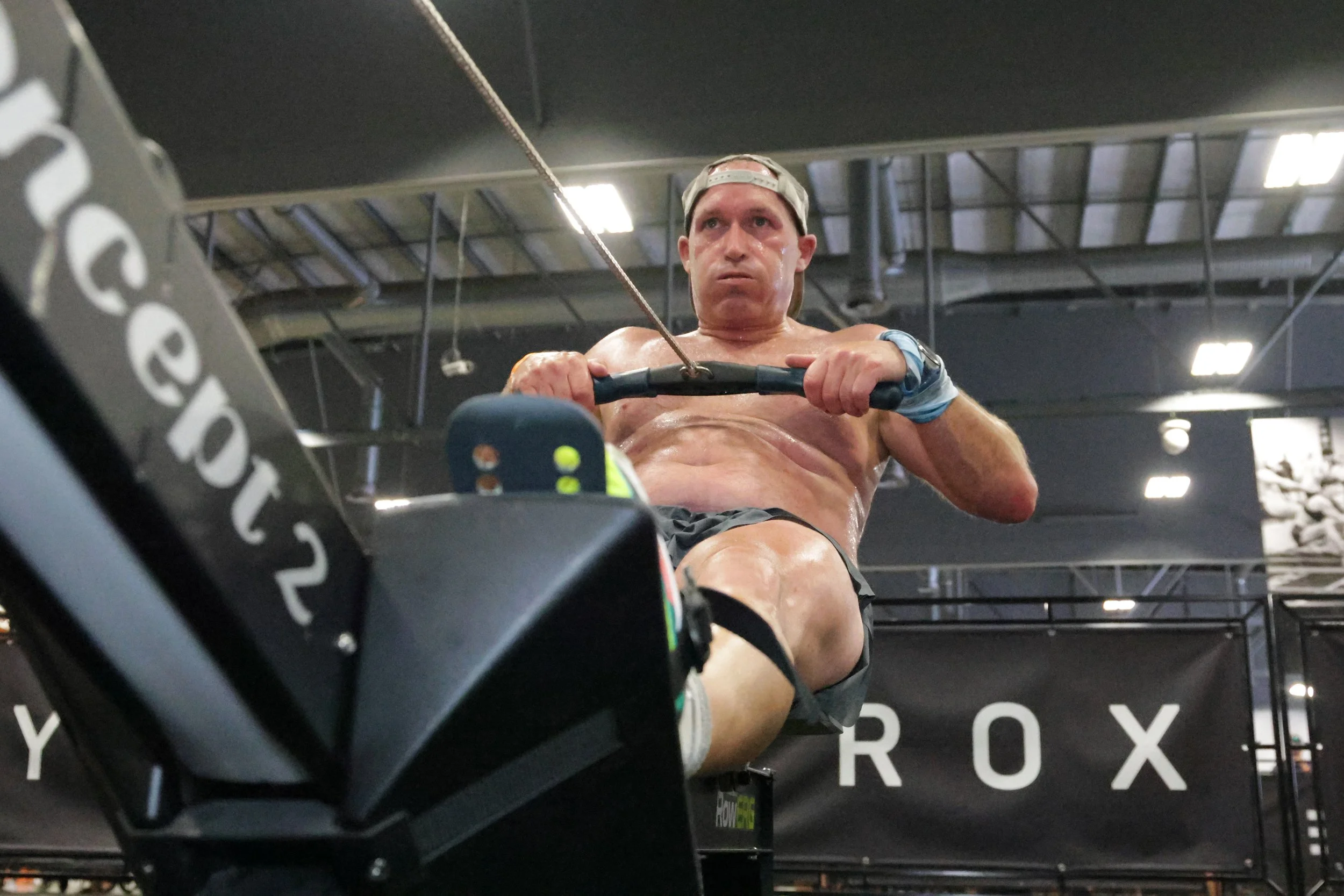 A shirtless man is working out on a rowing machine in a gym, with a focused expression, sweat visible on his face, and a baseball cap worn backwards. The gym has a black wall with the partial word 'ROX' visible in the background, and some overhead li