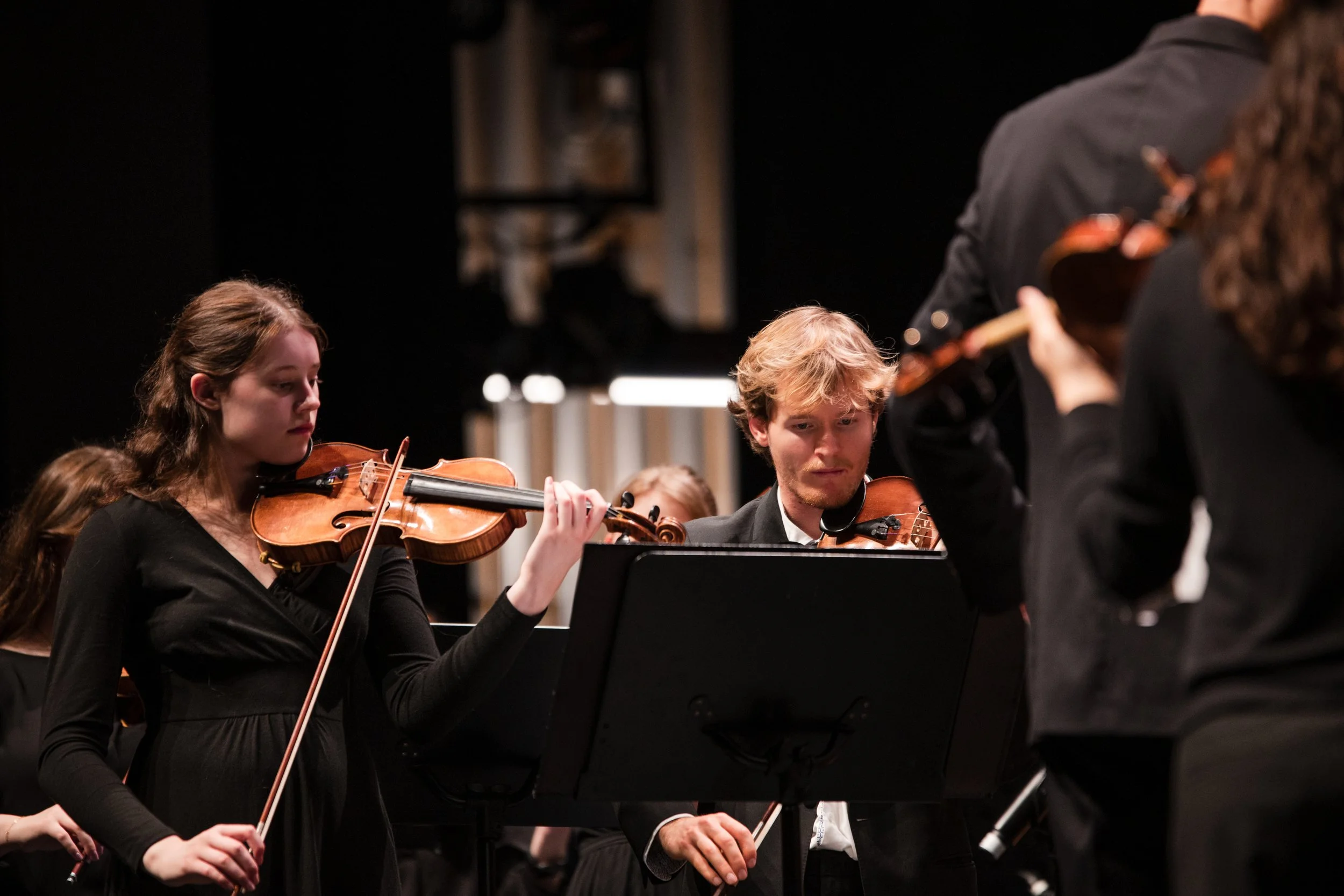 Orchestral musicians playing violins during a concert, focusing on two musicians, one female and one male, with a conductor partially visible in the right foreground.
