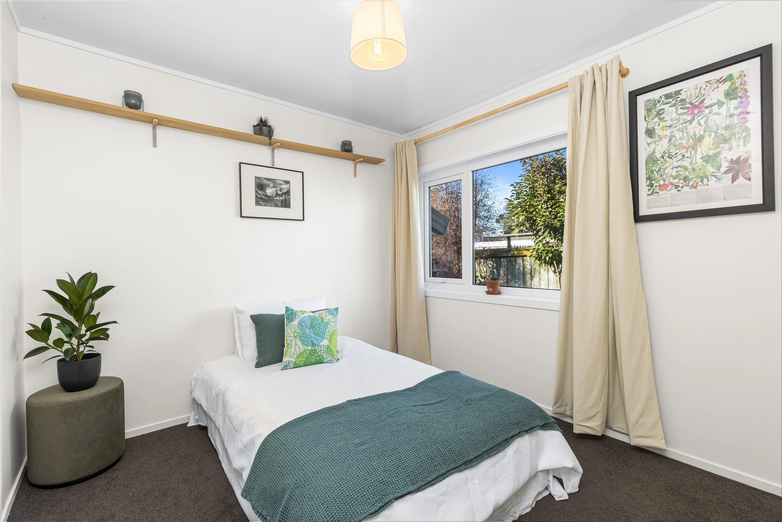 A bedroom with a bed featuring green and white pillows, a large window with beige curtains, framed artwork of botanical illustrations on the wall, a potted plant on a round side table, and a wooden shelf with small plants and decorative items, overal