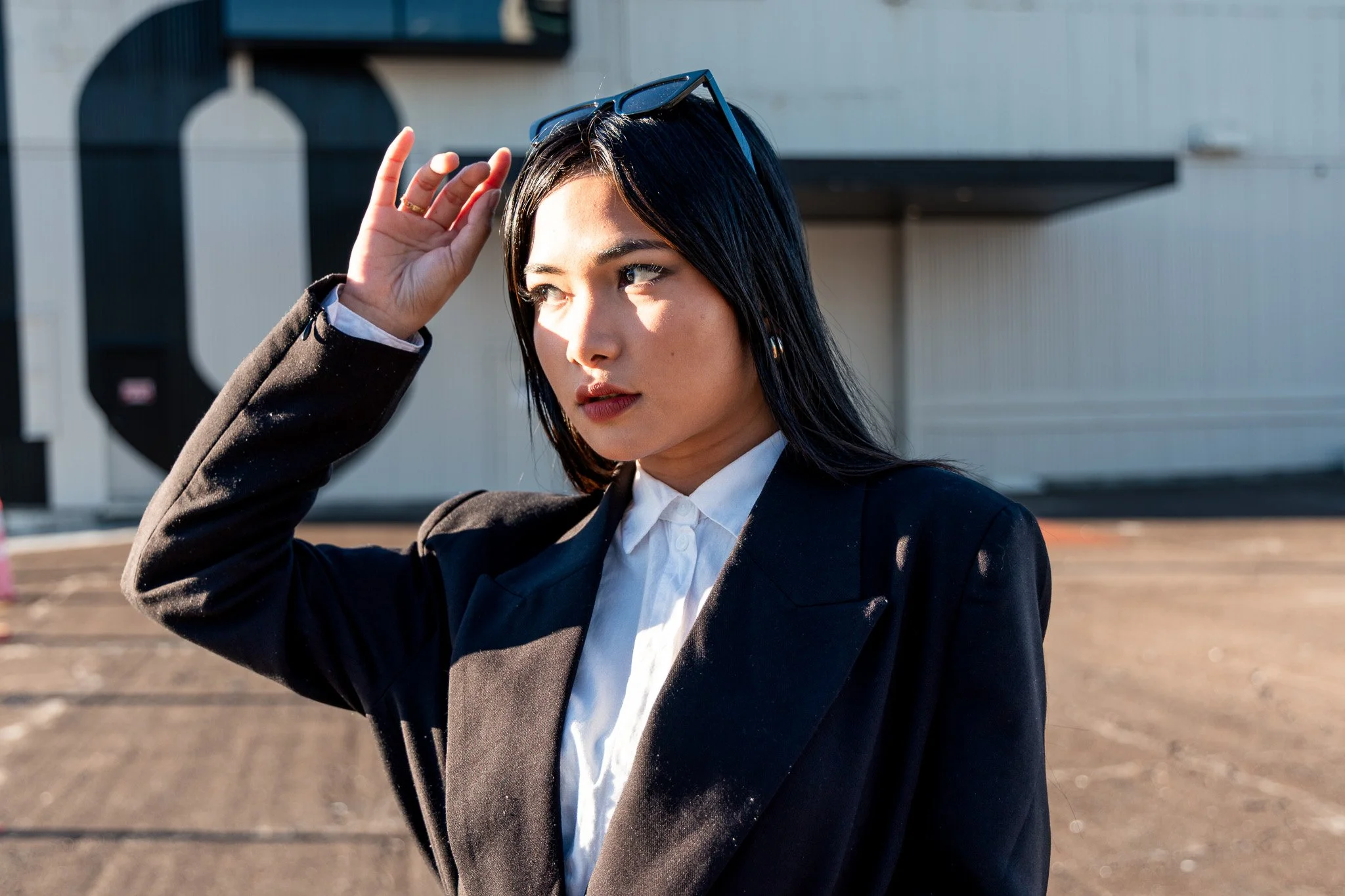 A young woman with black hair and light skin, dressed in formal business attire, adjusting her sunglasses outdoors on a sunny day.
