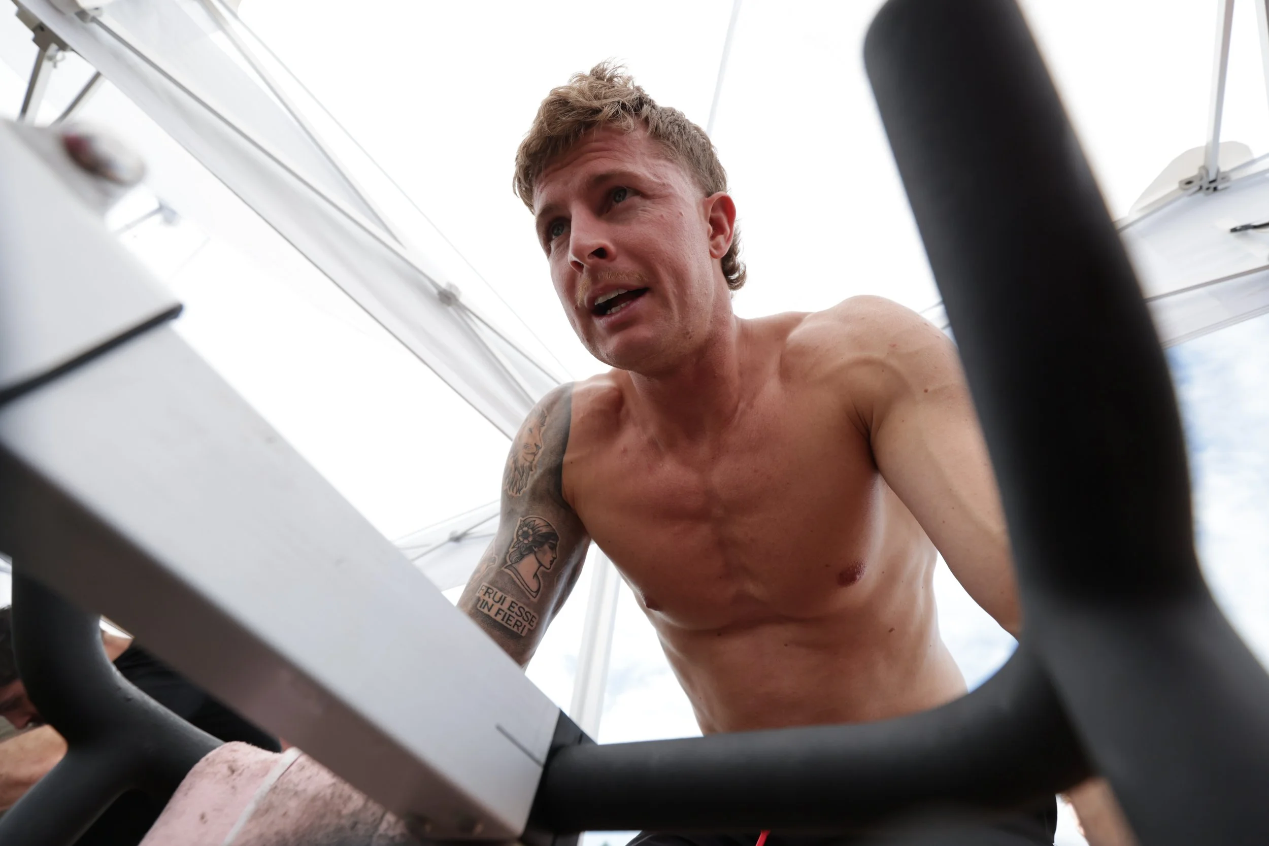 A shirtless man with tattoos on his arm is seen from below, looking down at the camera during a workout or fitness activity inside a bright room with white walls and ceiling.