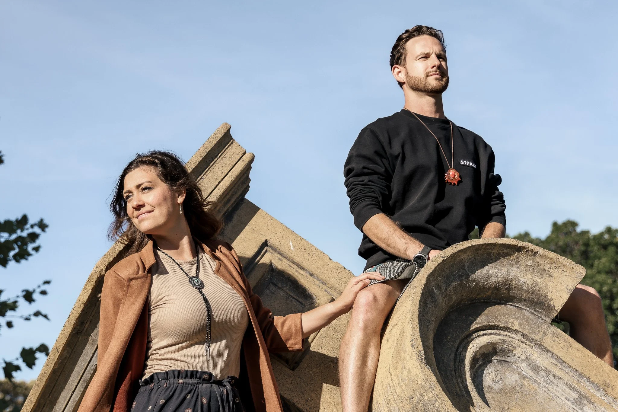 A man and woman sitting on a decorative stone structure outdoors, with a clear blue sky and trees in the background.