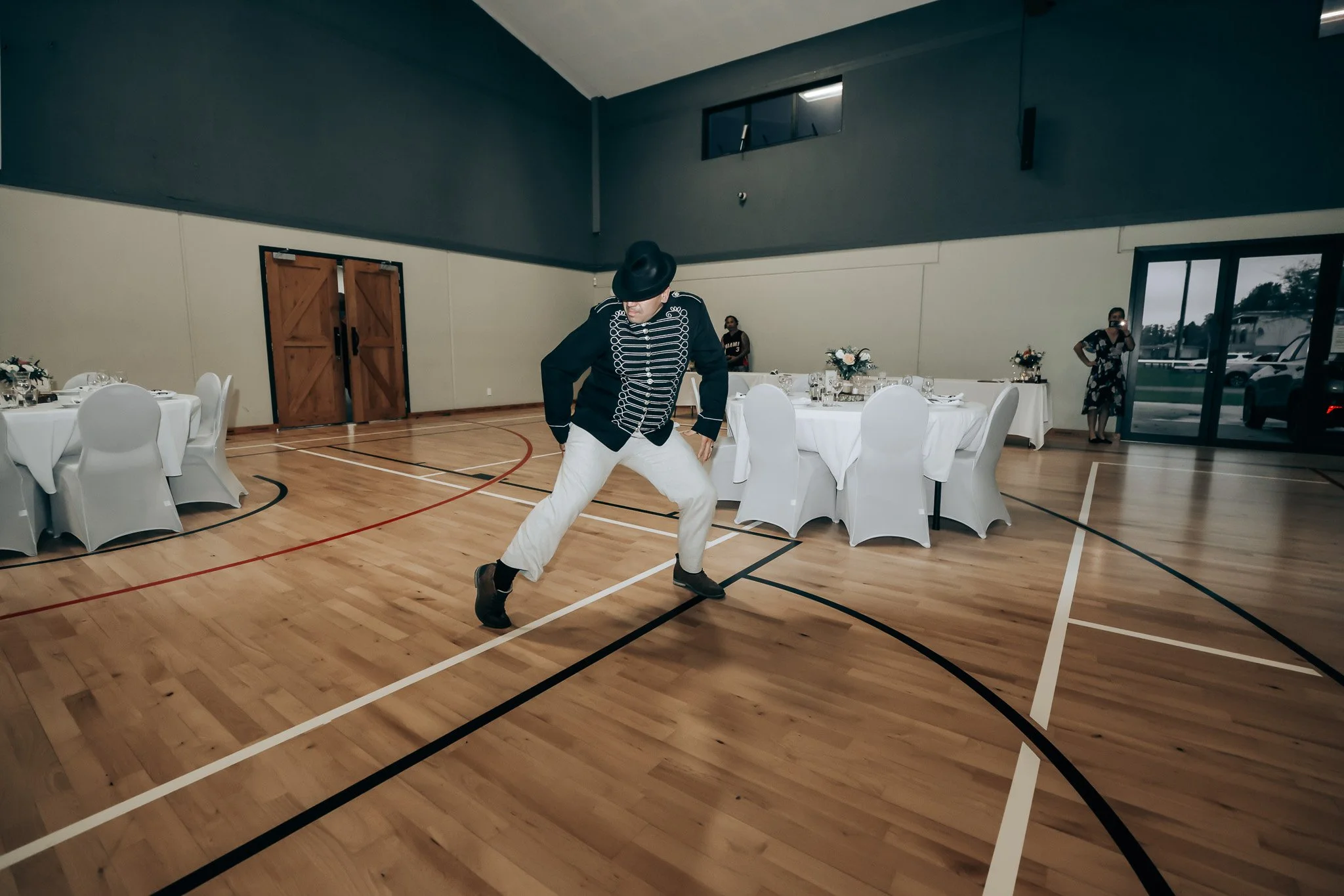 A person wearing a black hat, black jacket with white decorative lines, and light-colored pants is performing a dance move in a gymnasium with wooden floors and round banquet tables covered with white tablecloths, decorated with flower centerpieces. 
