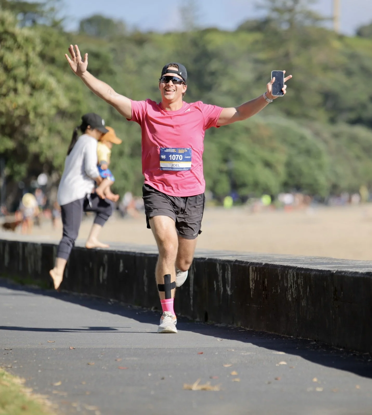 A man running in a race, wearing a pink shirt, black shorts, pink socks, a prosthetic leg, and sunglasses, holding a phone in his right hand, with an event bib number 1070. He is smiling and has his arms raised in celebration. In the background, ther
