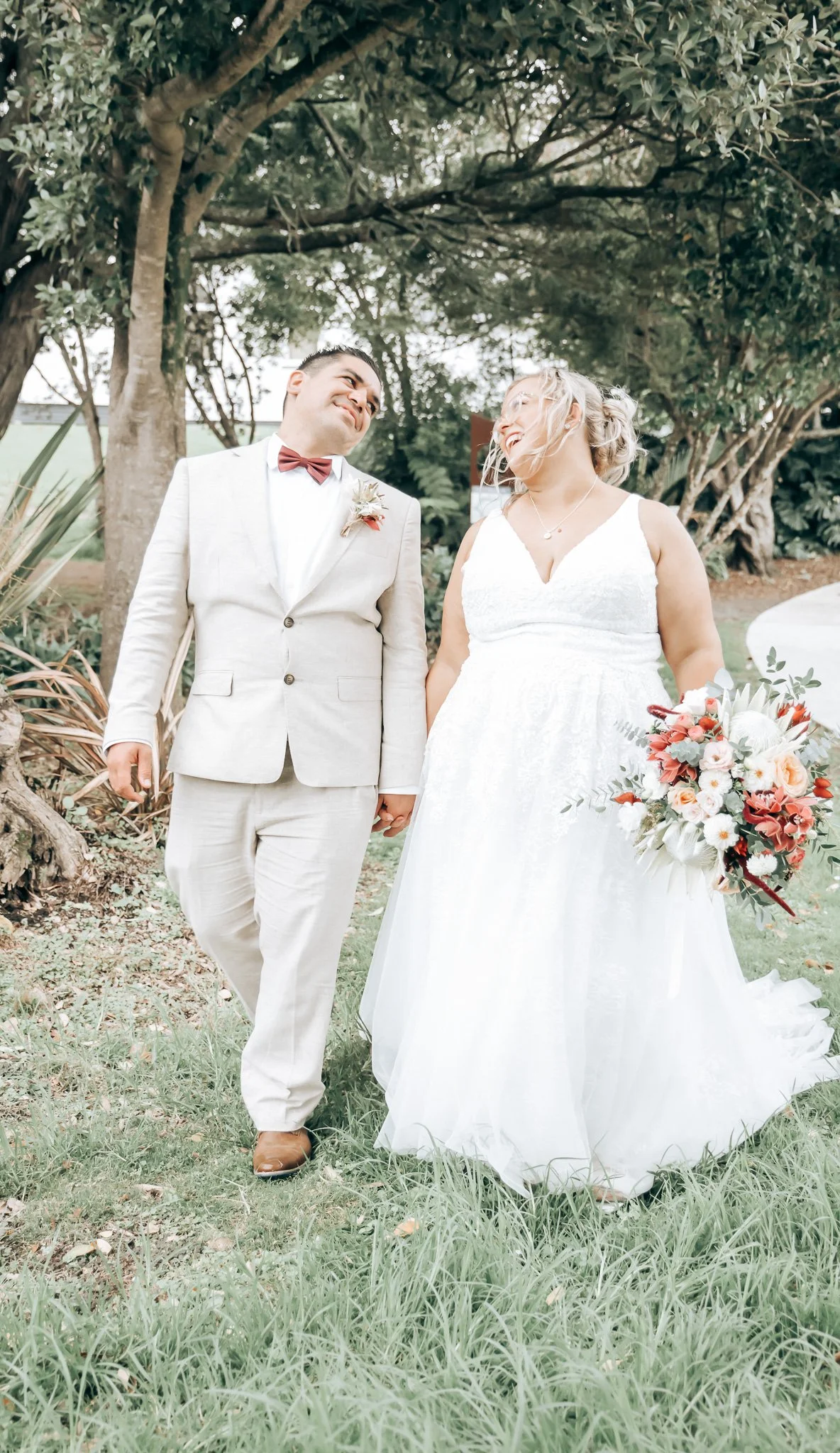 A newlywed couple standing hand in hand outdoors, smiling and looking at each other, with green trees and grass in the background.