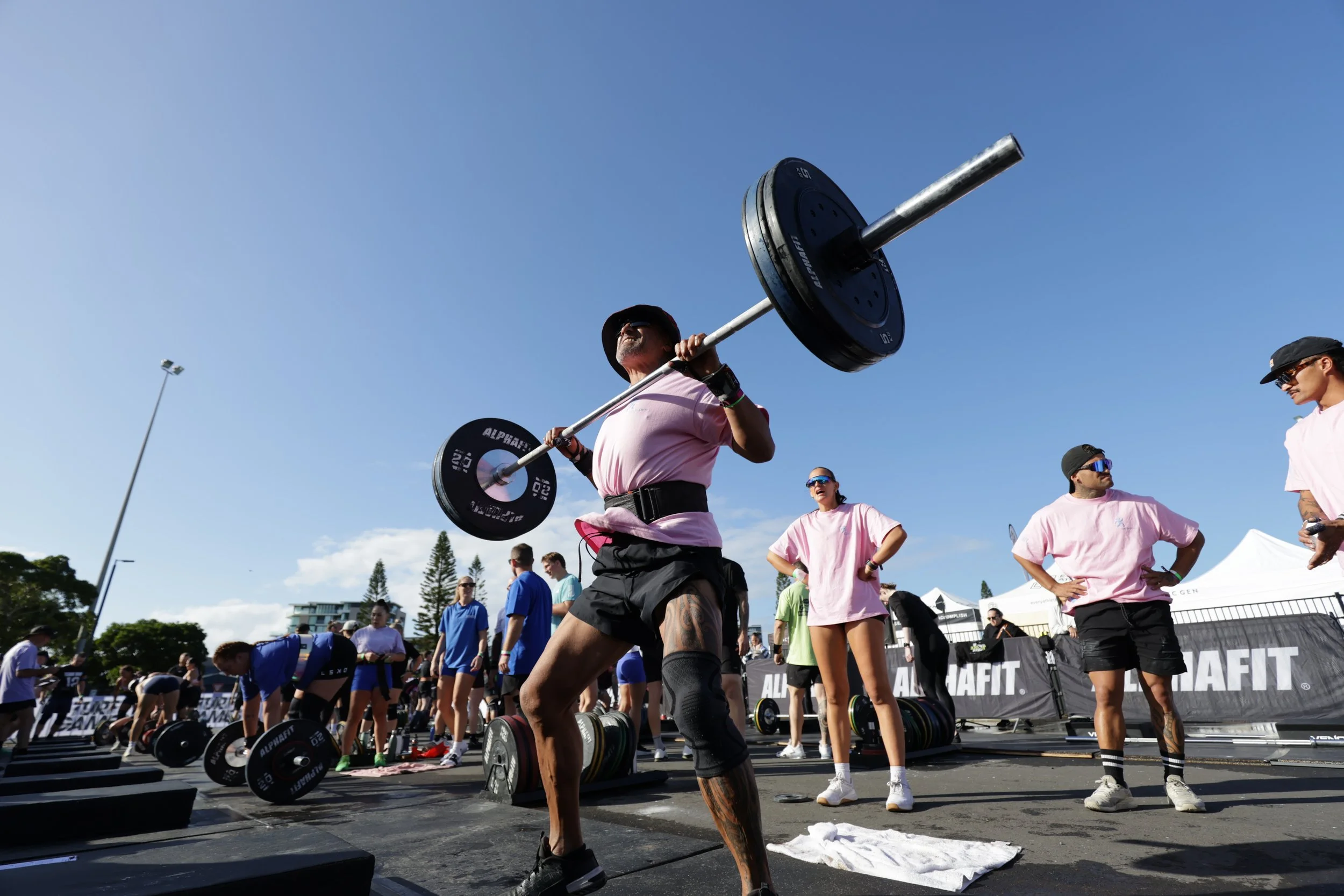Group of people participating in an outdoor weightlifting event, with a man in the foreground lifting a barbell overhead against a blue sky.