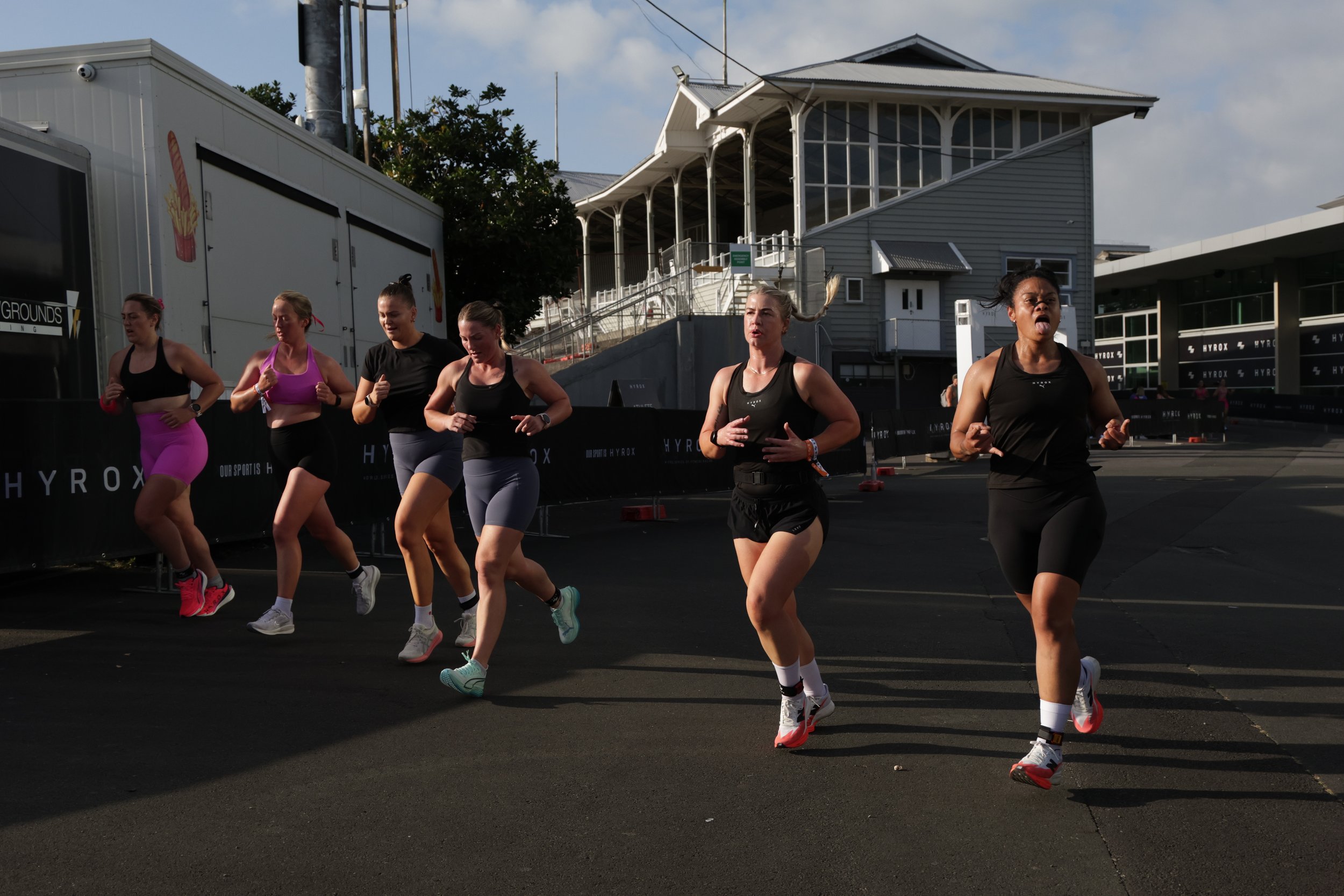 Group of women running outdoors during a race, dressed in athletic wear, with a building and cloudy sky in the background.