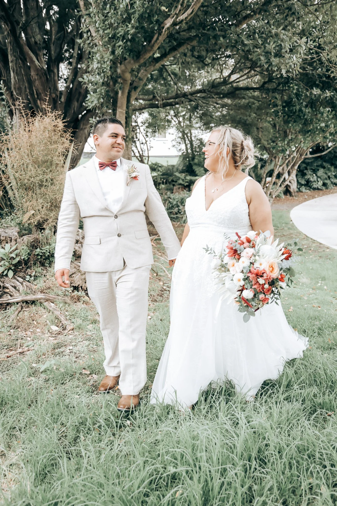 A bride and groom walking hand in hand through a park or garden, surrounded by greenery and trees, during their wedding day.