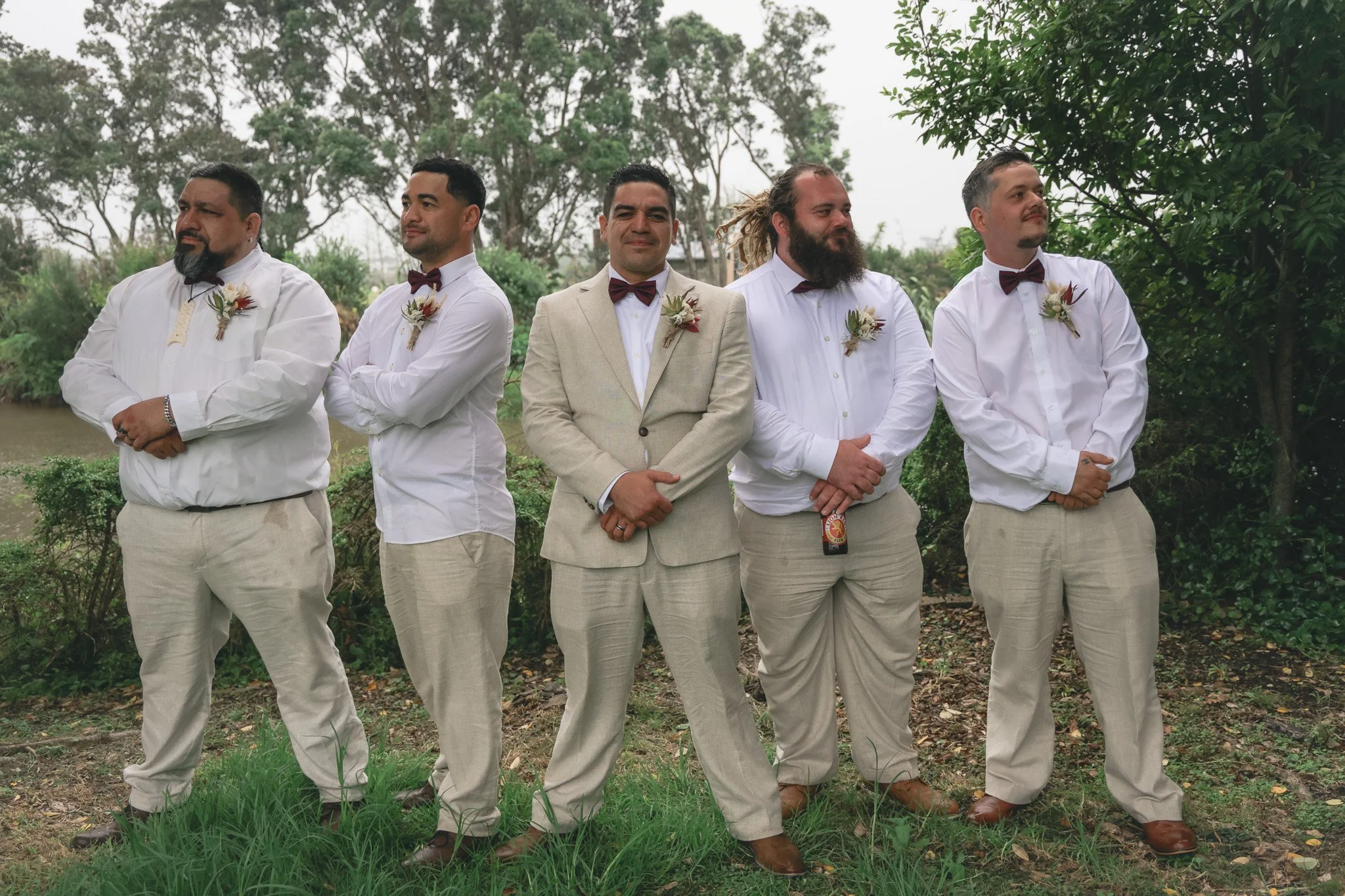 Five men standing outdoors in a line, dressed in formal attire with white shirts, beige pants, and maroon bow ties, with boutonnières on their shirts, on a grassy area with trees in the background.