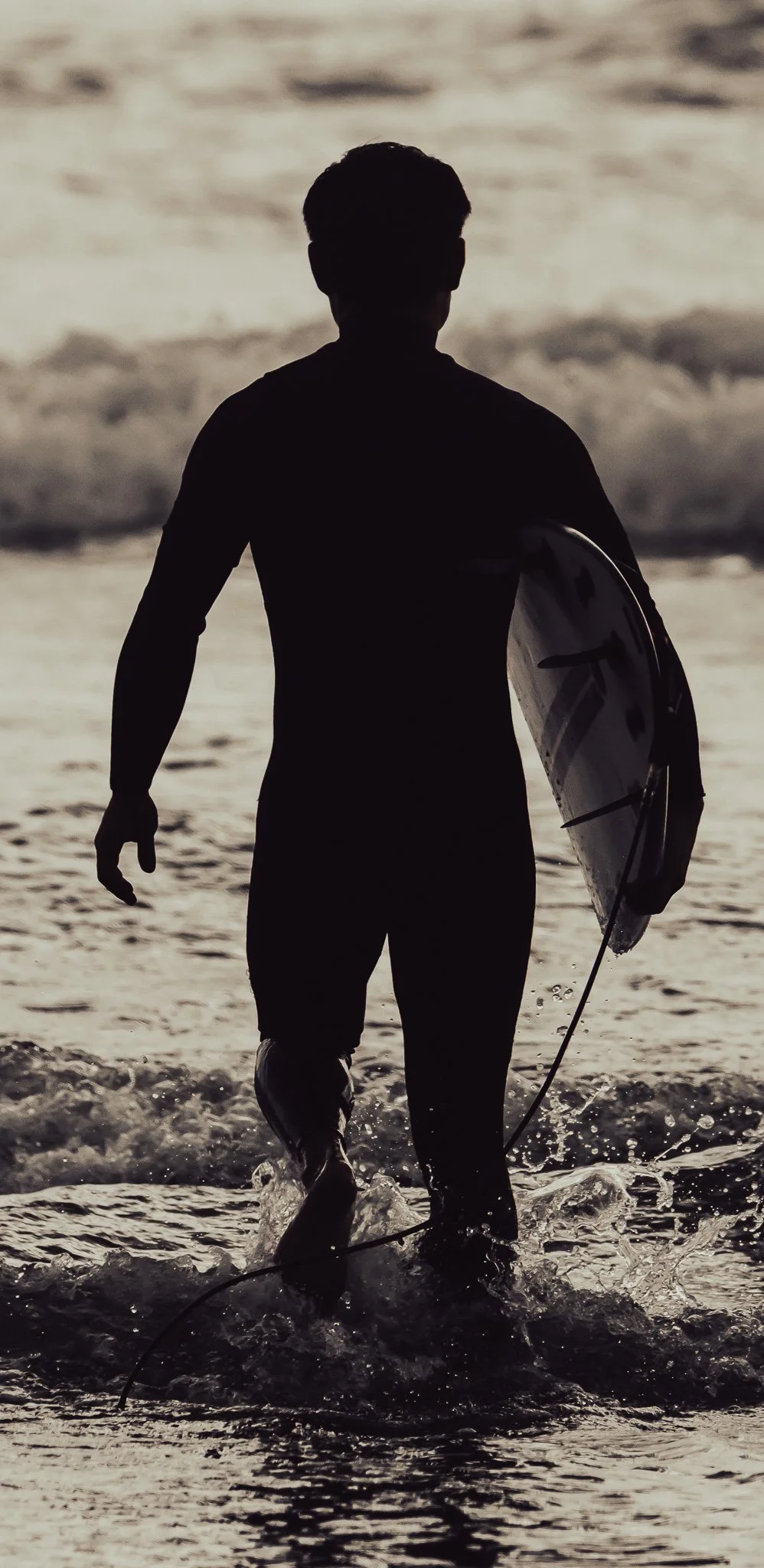 Silhouette of a man walking out of the ocean holding a surfboard during sunset.