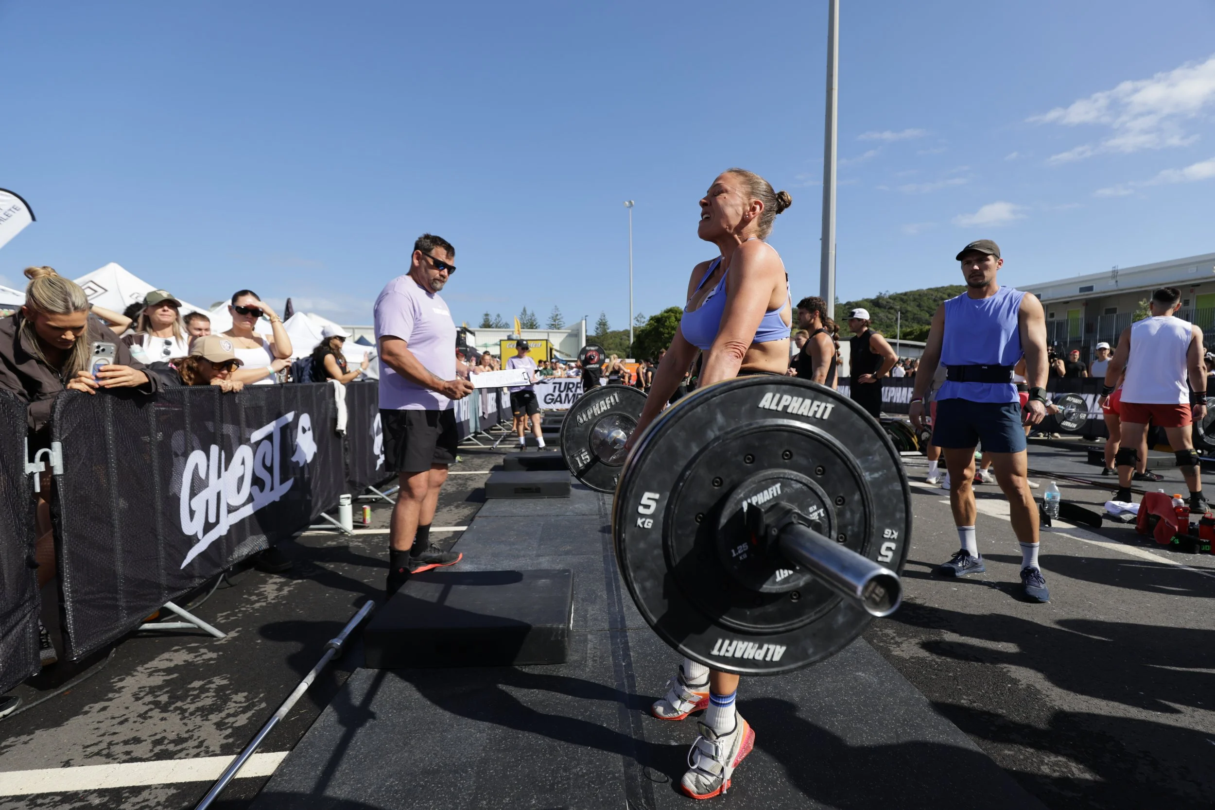 A woman lifting a barbell during a CrossFit competition outdoors on a sunny day, with spectators and other athletes watching.