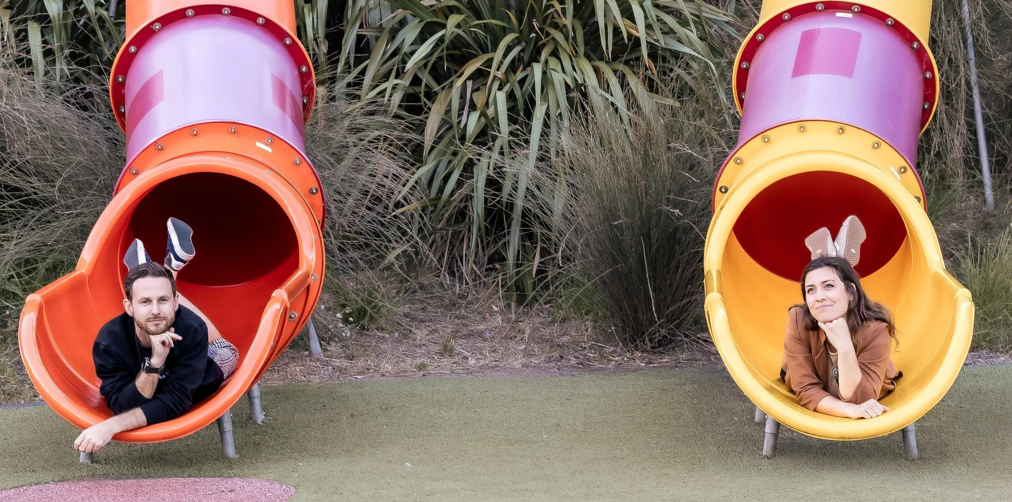 Two people lying on the ground with their heads and shoulders inside colorful tube slides at a playground. The man on the left has bunny ears and is resting on his hand, while the woman on the right has bunny ears and is resting her chin on her hand,