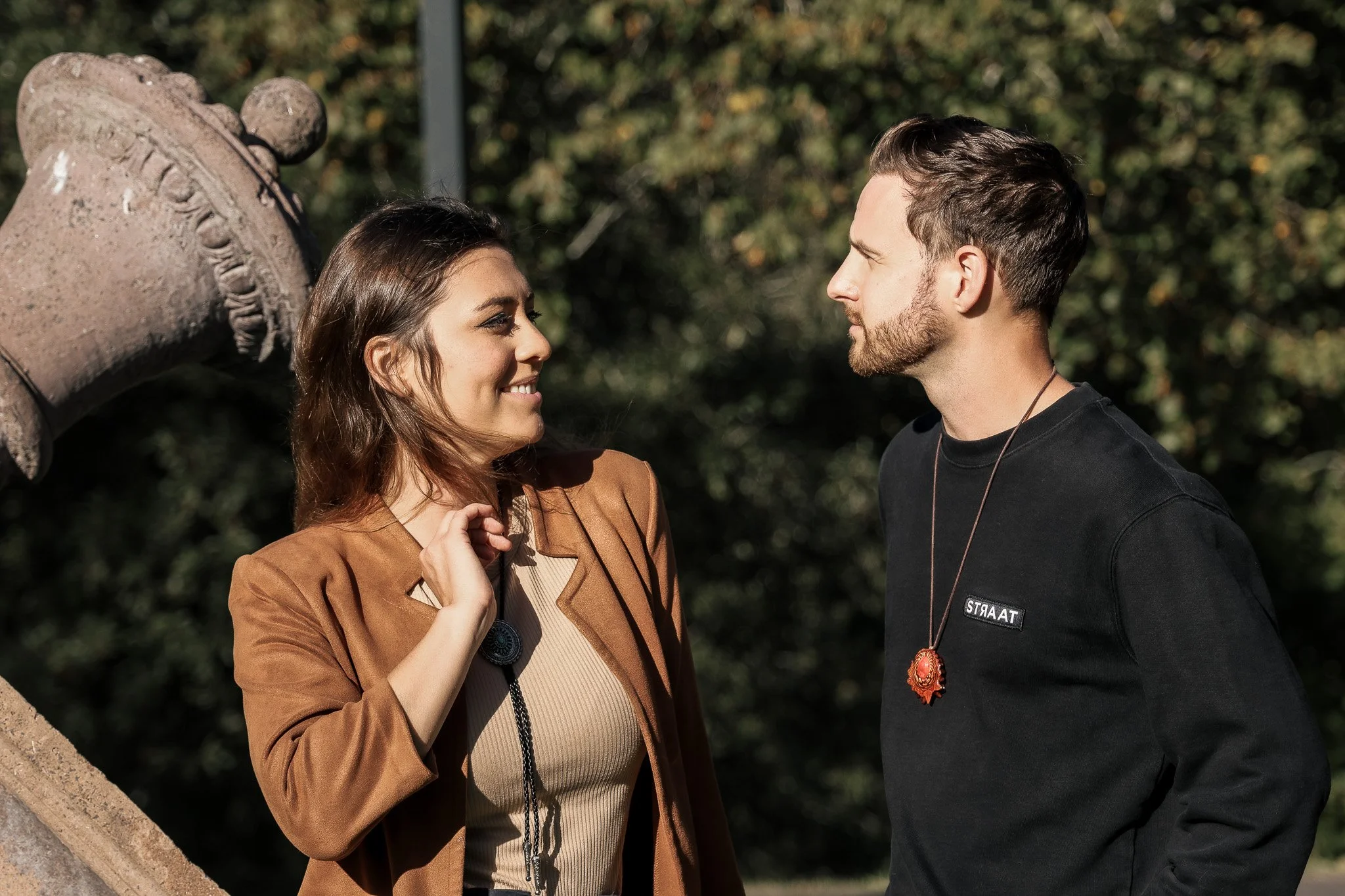 A young woman with brown hair smiling and a young man with brown hair and a beard standing face to face outdoors near a stone structure with trees in the background.